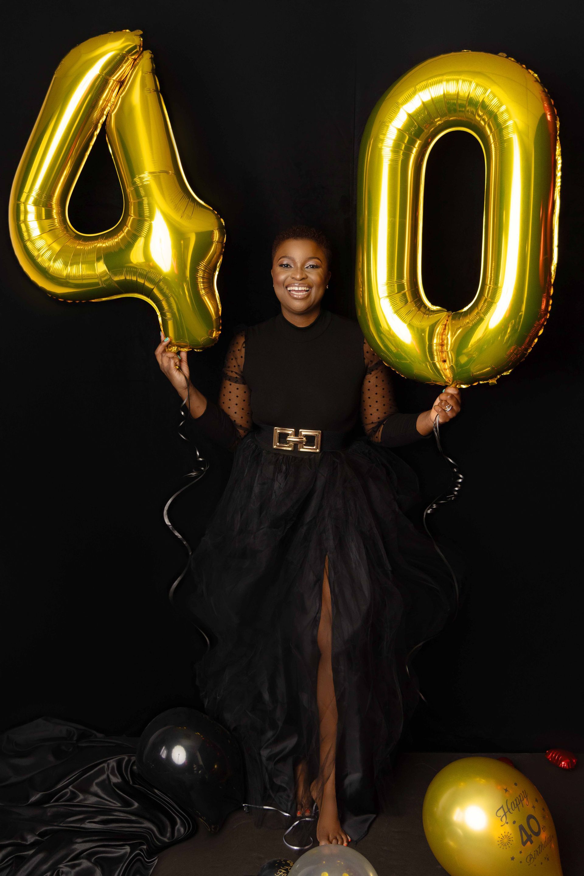 A woman in a black dress is holding balloons in the shape of the number 40.