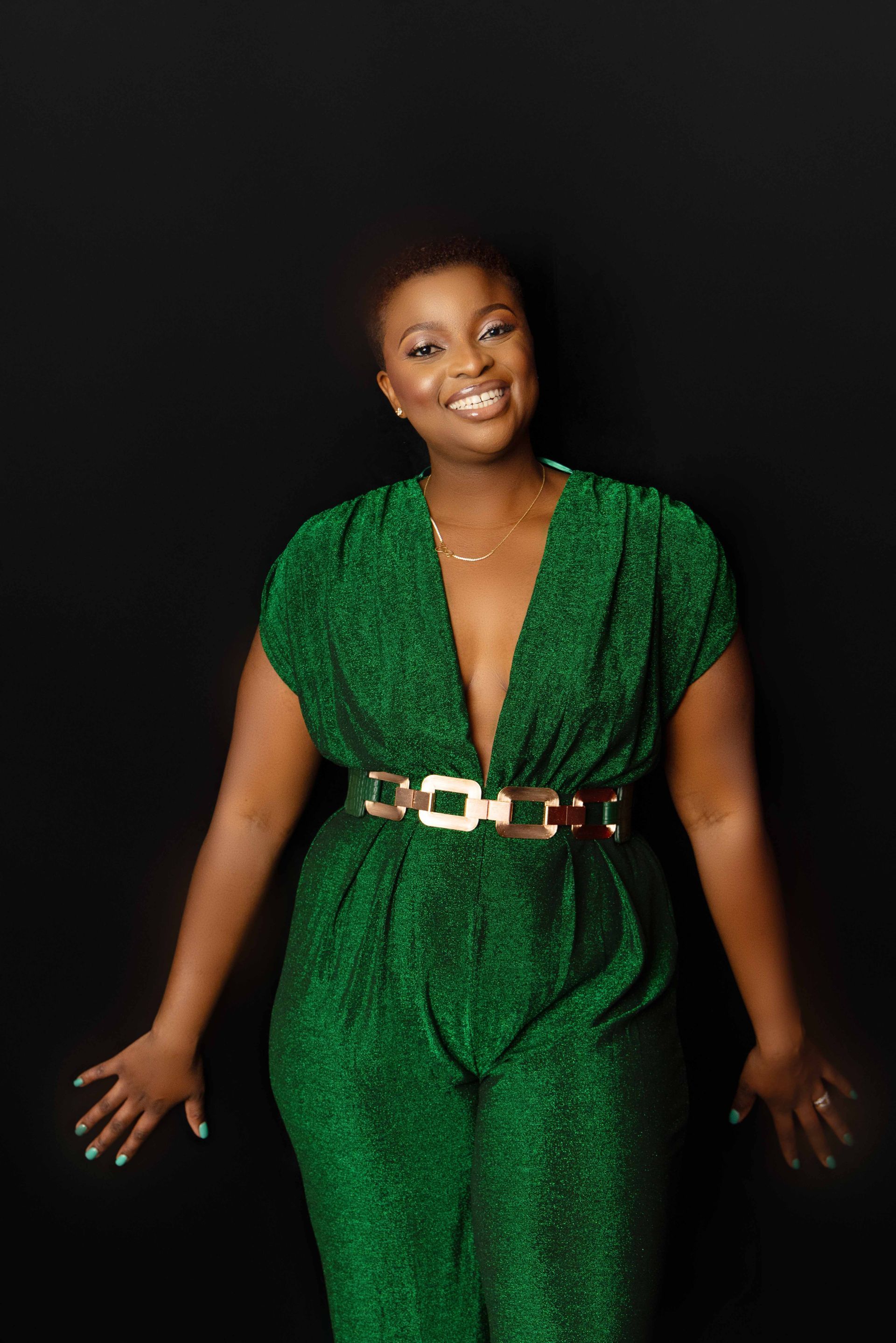 A woman in a green jumpsuit is standing in front of a black background.