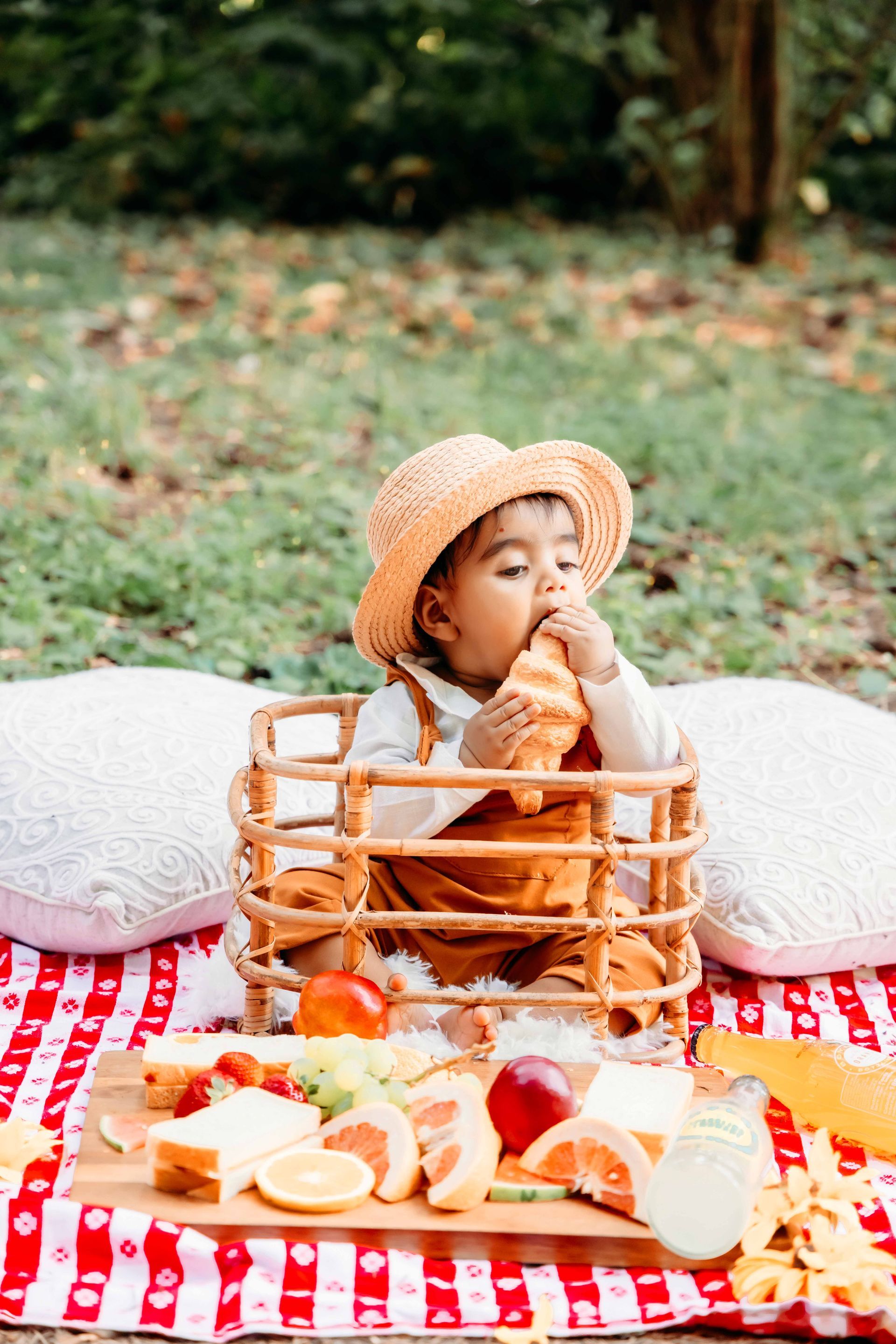 A baby is sitting on a picnic blanket eating a croissant.