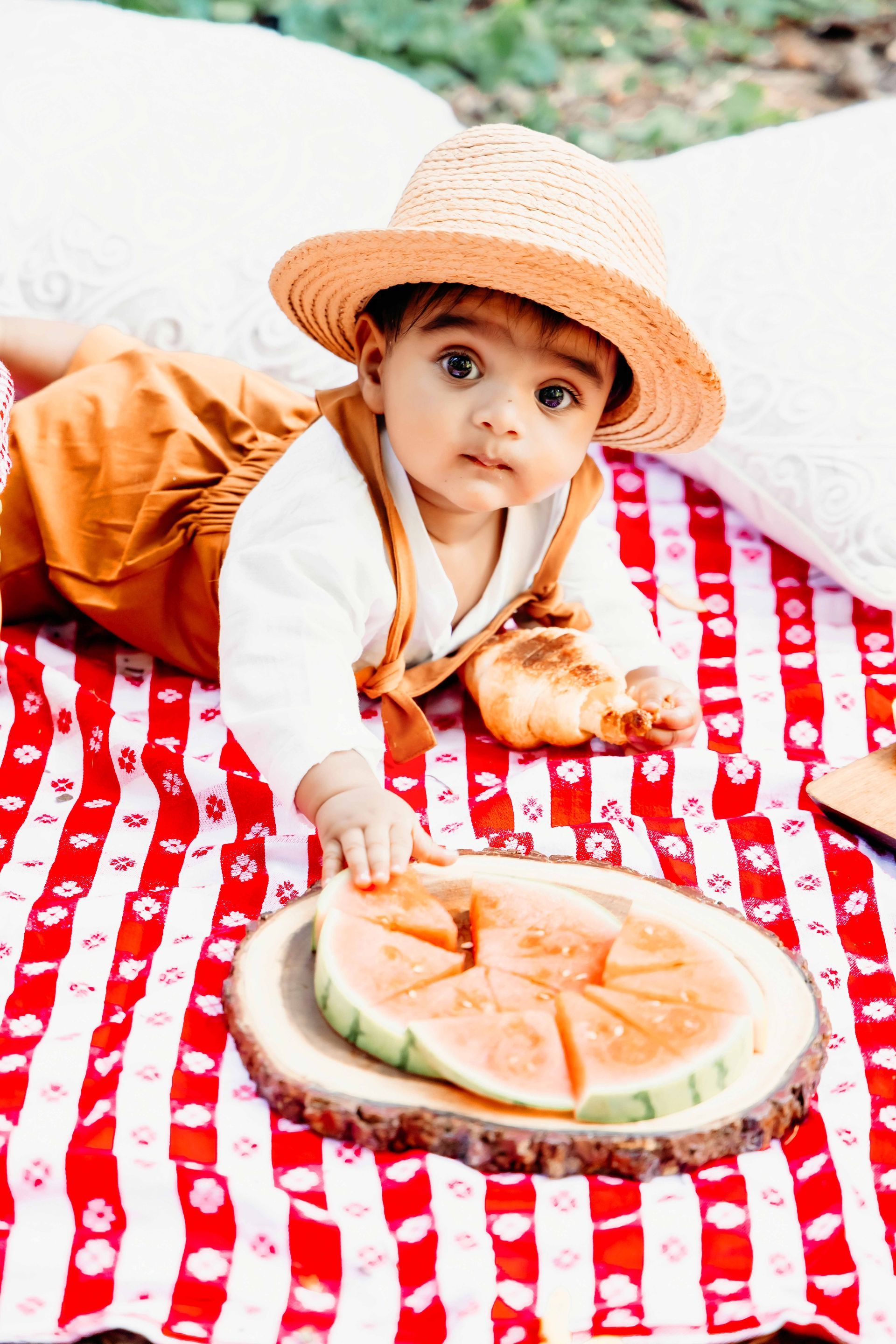 A baby is laying on a picnic blanket with a slice of watermelon.