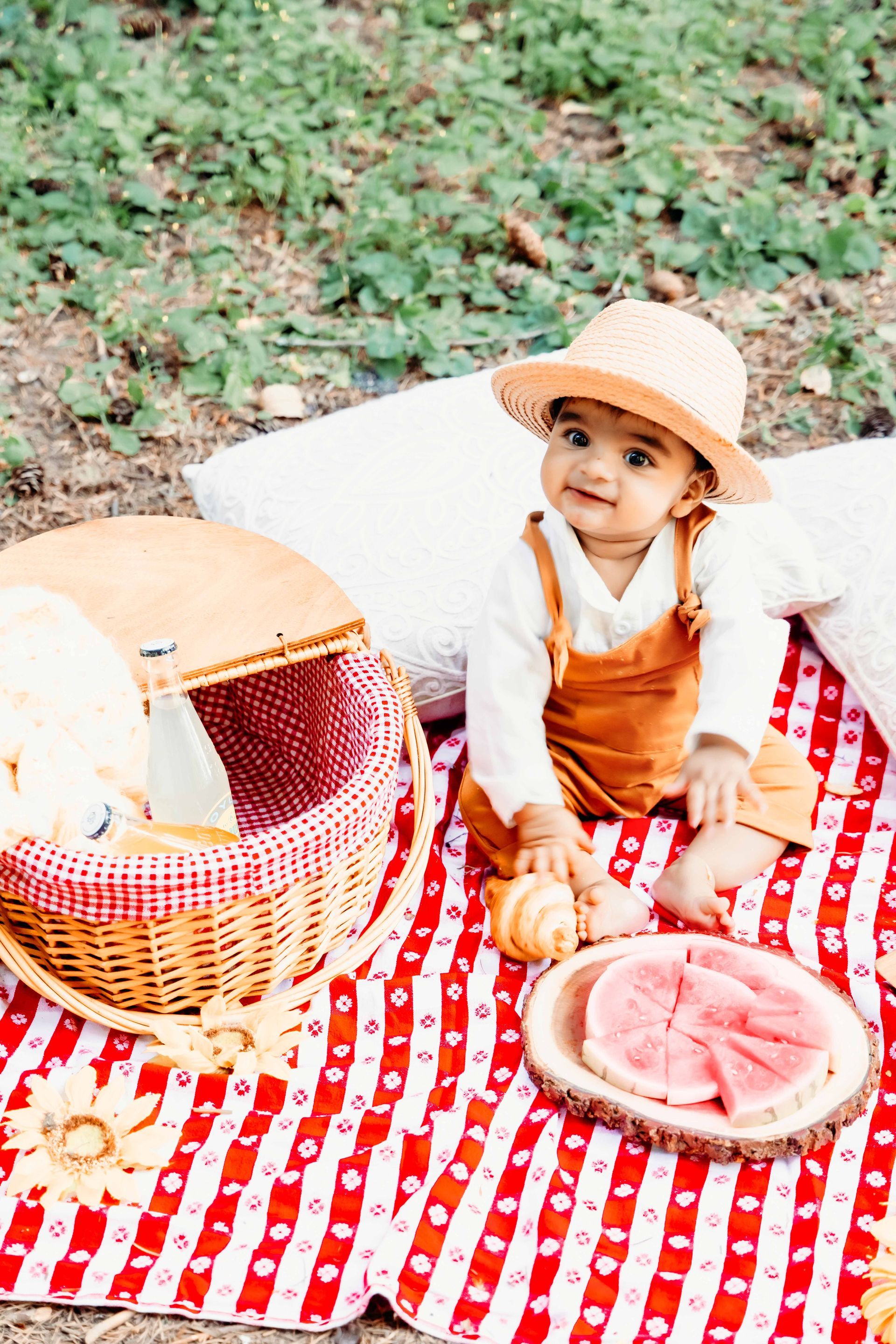 A baby is sitting on a picnic blanket with a basket and watermelon.