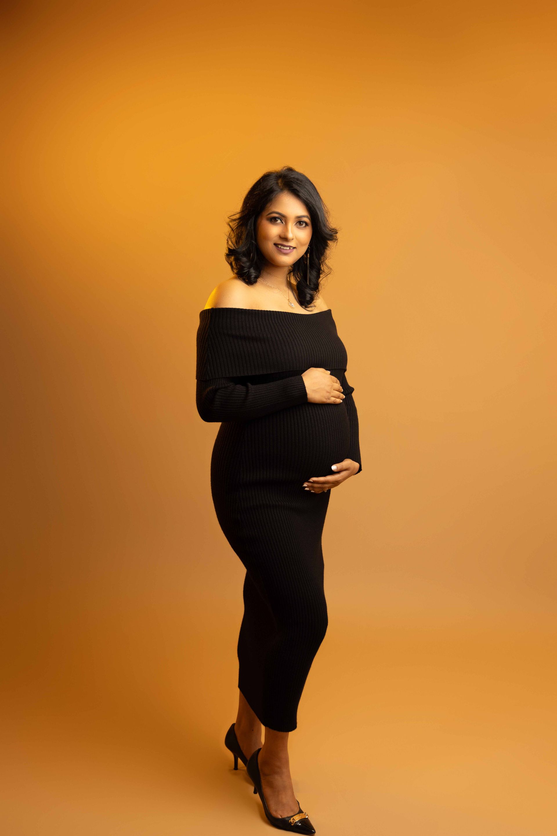 A pregnant woman in a black dress is standing in front of a yellow background.