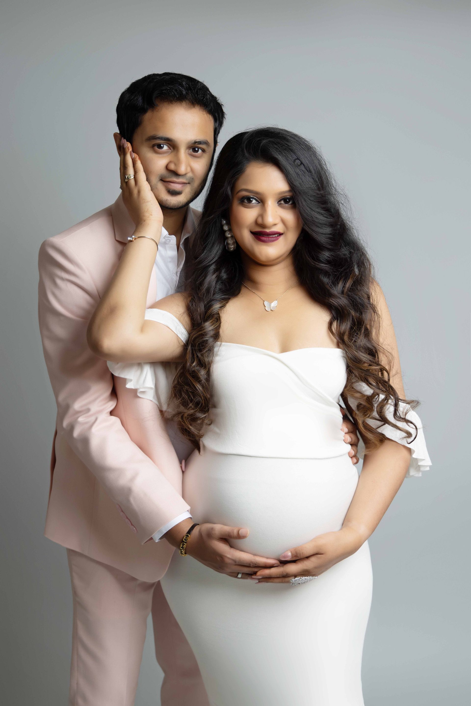 A man in a pink suit is hugging a pregnant woman in a white dress.