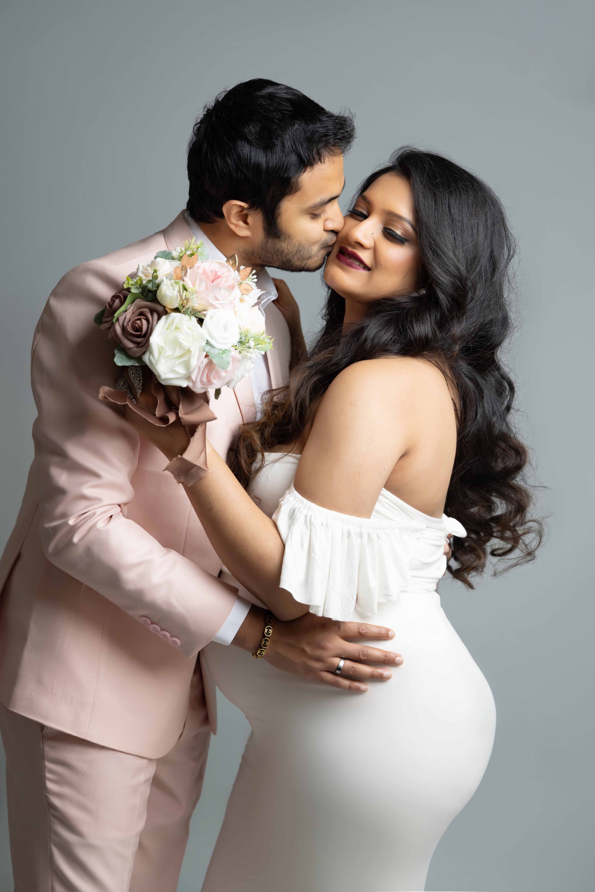 A man is kissing a pregnant woman on the cheek while she holds a bouquet of flowers.