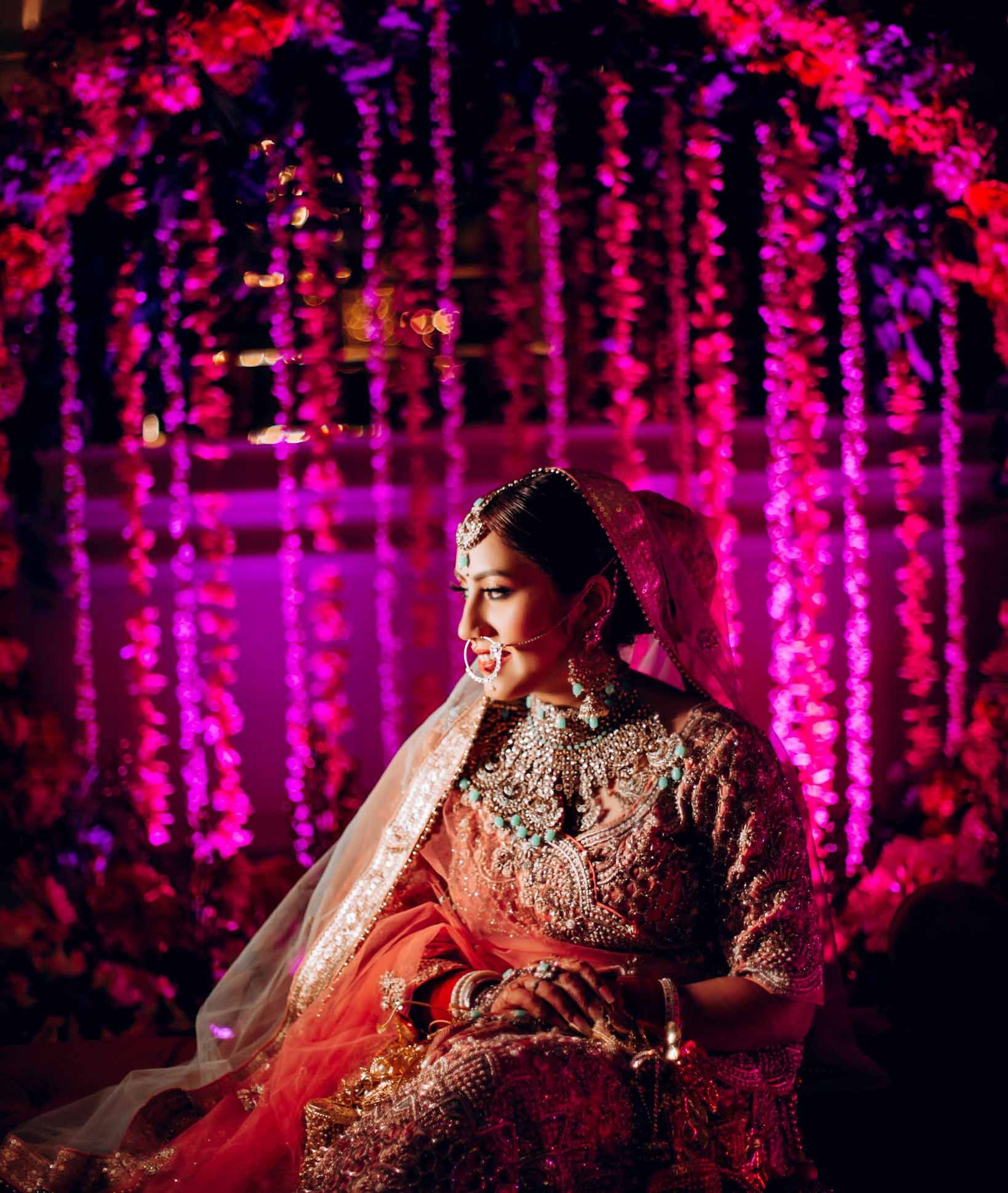 A bride in a red and gold dress is sitting in front of a pink background.