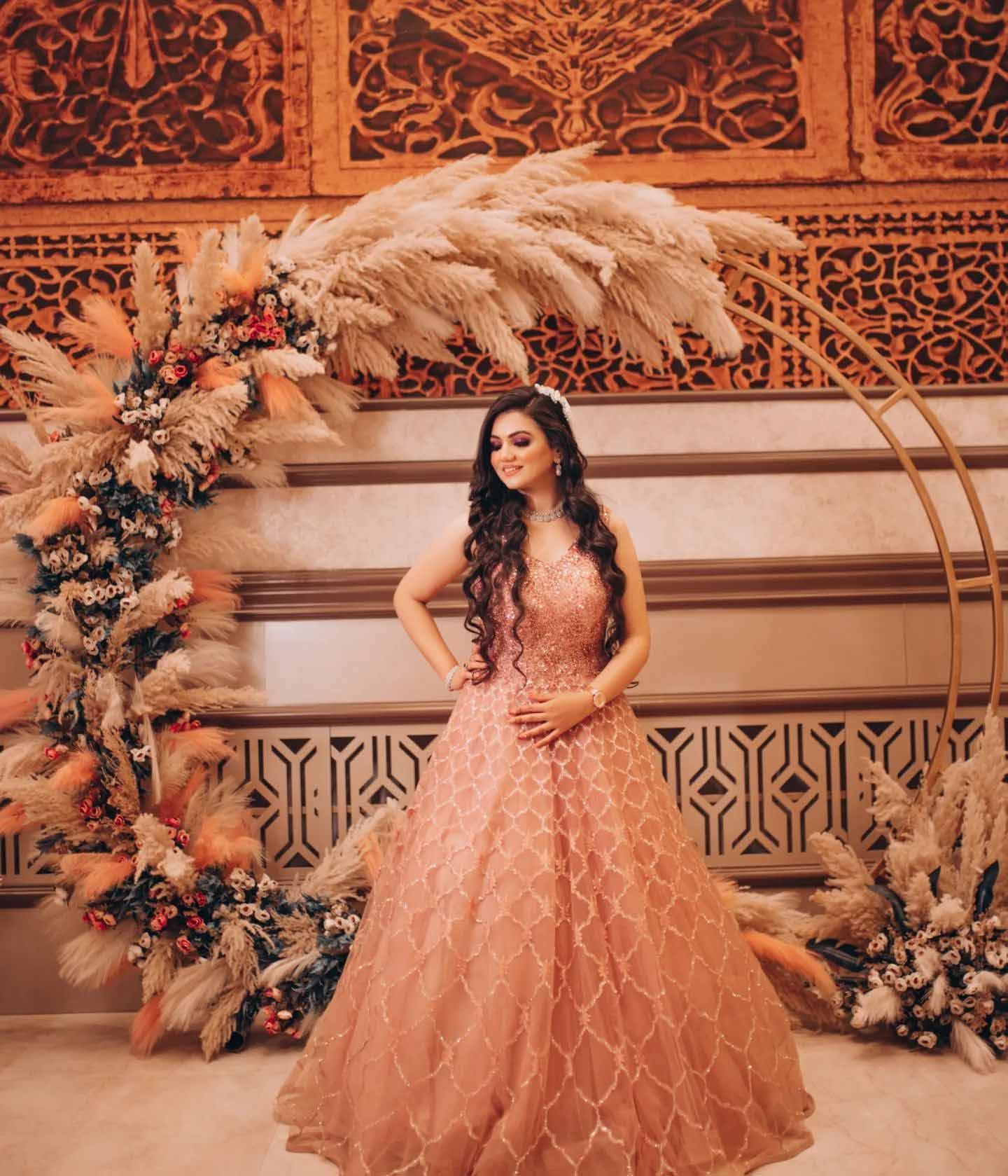A woman in a long pink dress is standing in front of a floral arch.