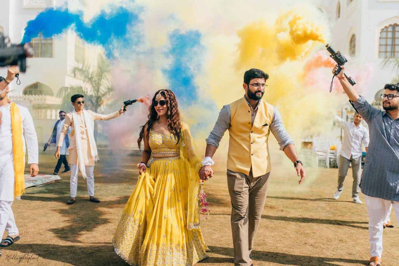 A bride and groom are walking through a crowd of people holding colored smoke.