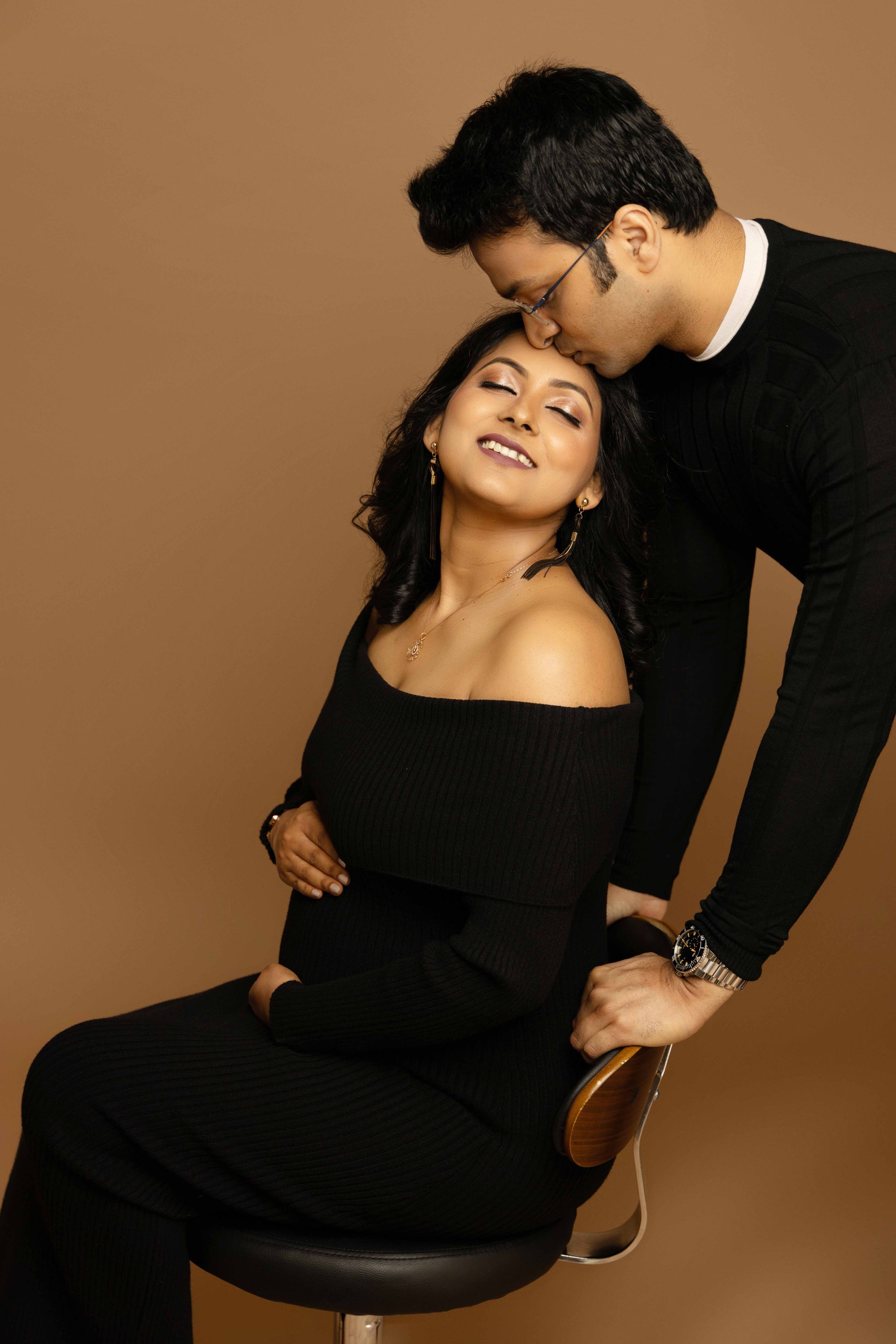 A man is kissing a pregnant woman who is sitting on a chair.