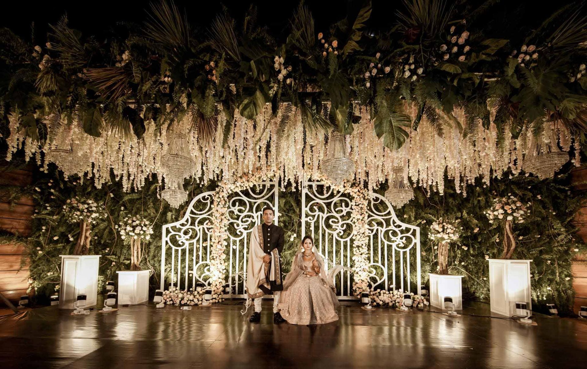 A bride and groom are standing in front of a stage decorated with flowers and lights.