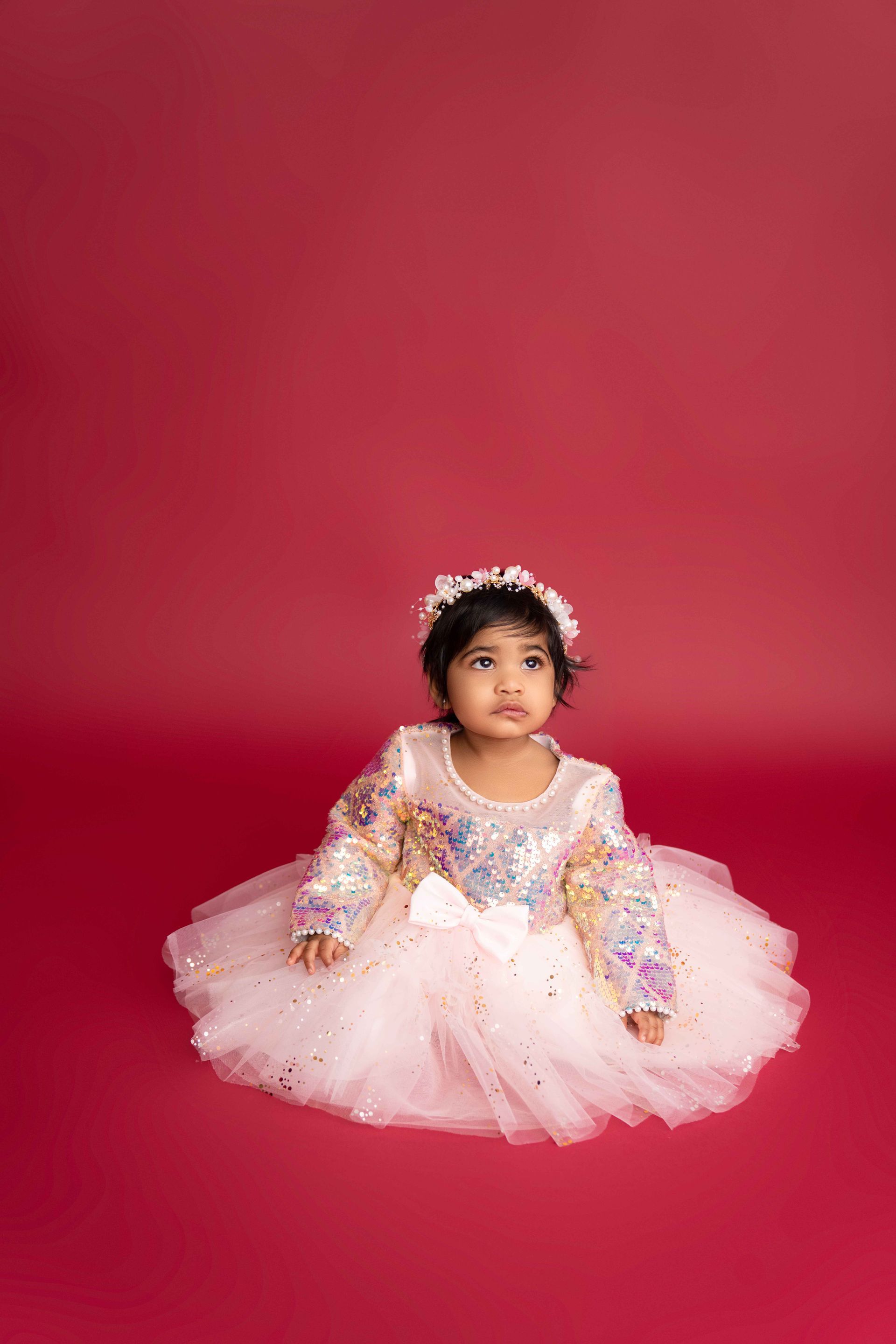 A little girl is sitting on the floor wearing a white dress and a flower crown.
