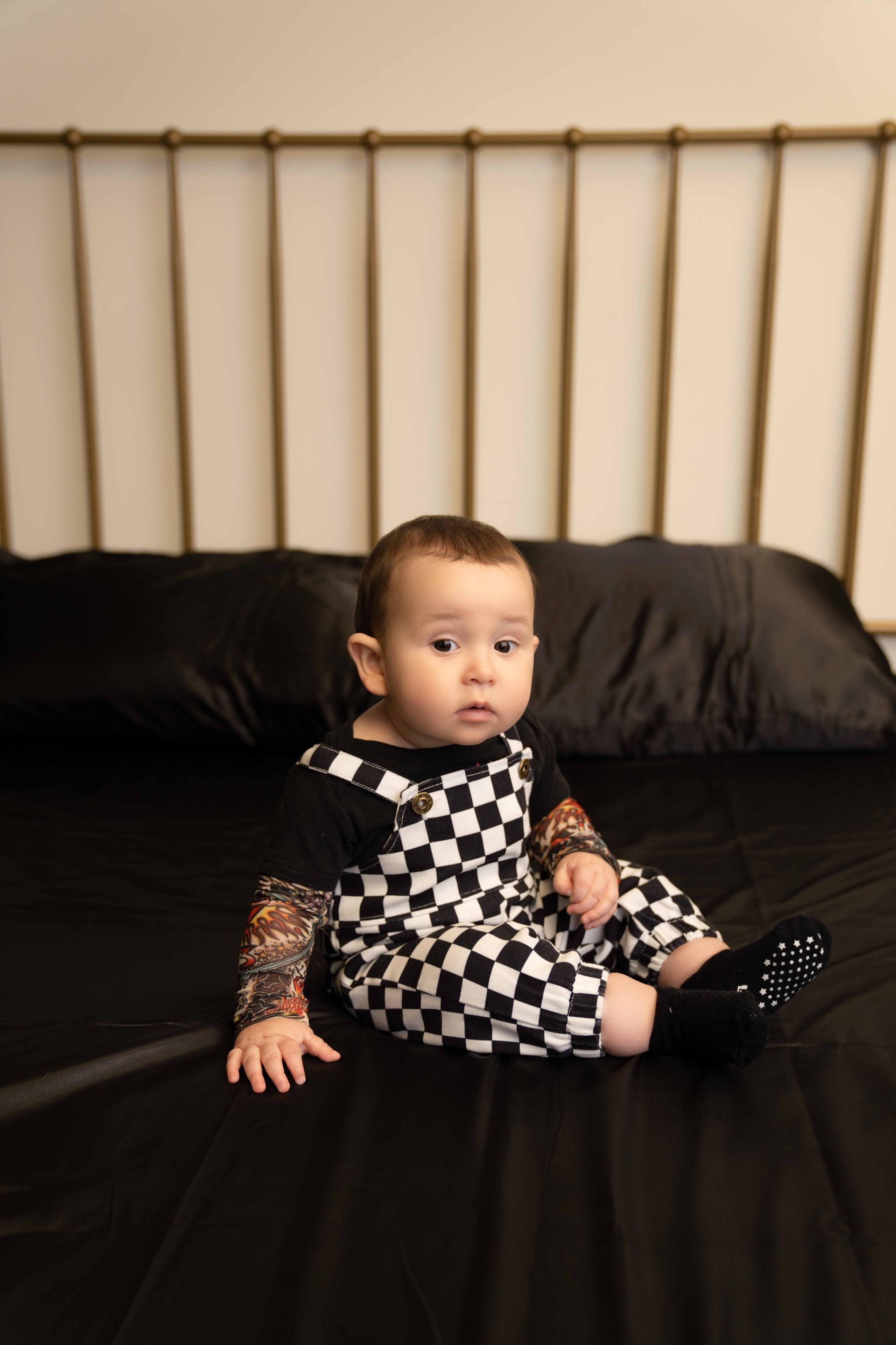 A baby is sitting on a bed wearing black and white checkered overalls.
