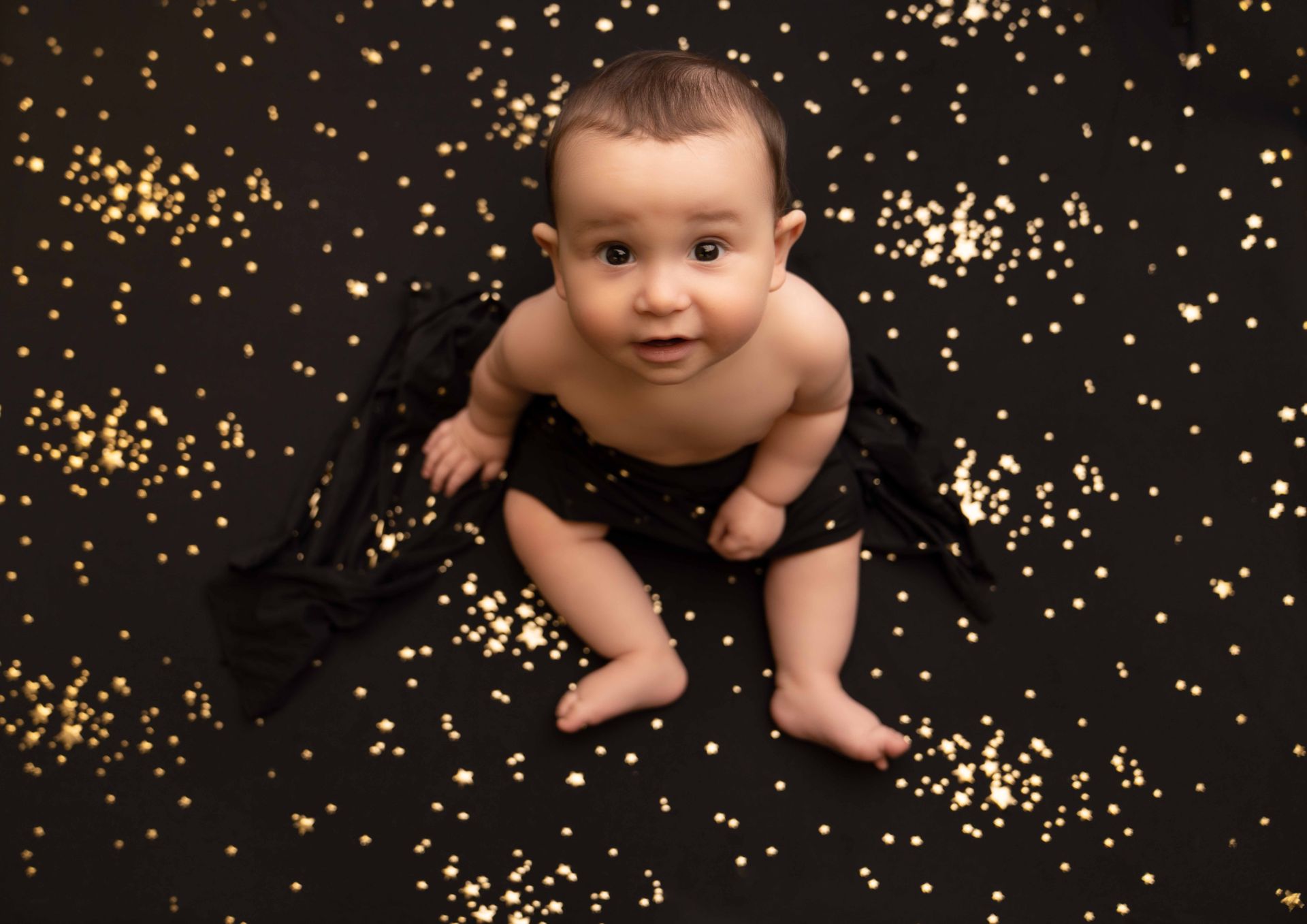 A baby is sitting on a black blanket with gold stars.