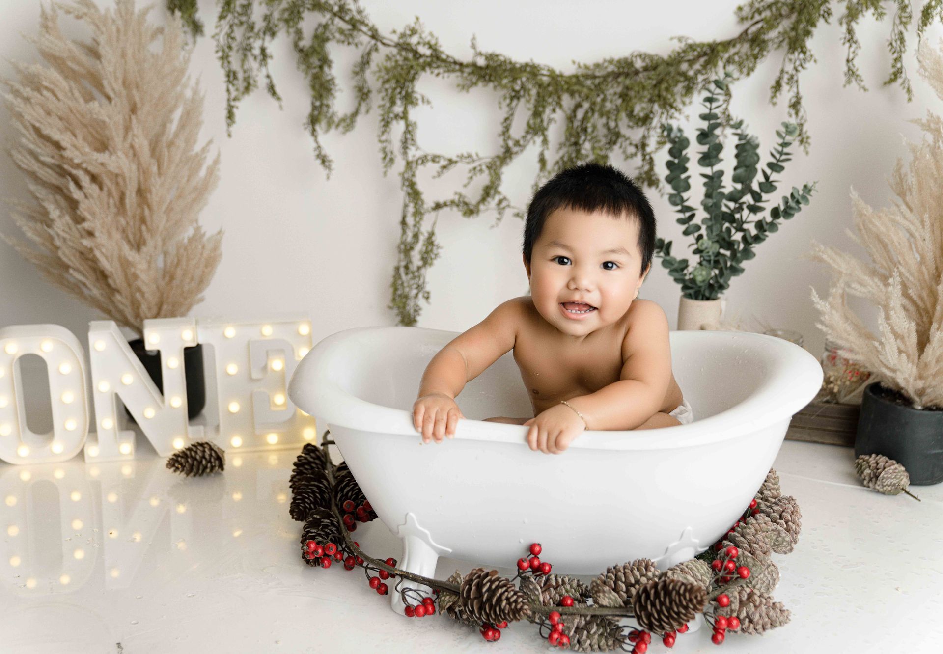 A baby is sitting in a bathtub decorated with pine cones and berries.