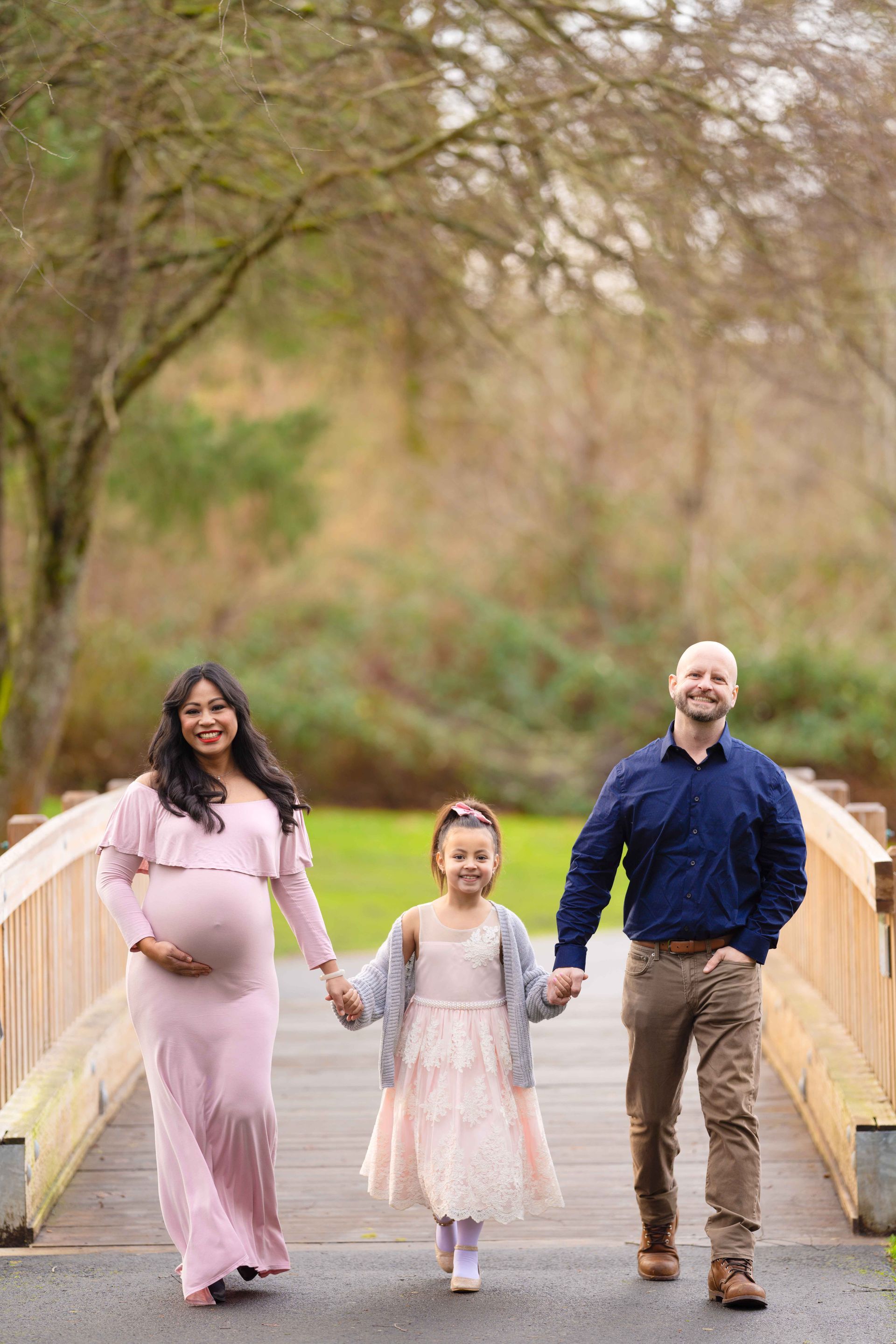 A pregnant woman, a man and a little girl are standing on a bridge holding hands.