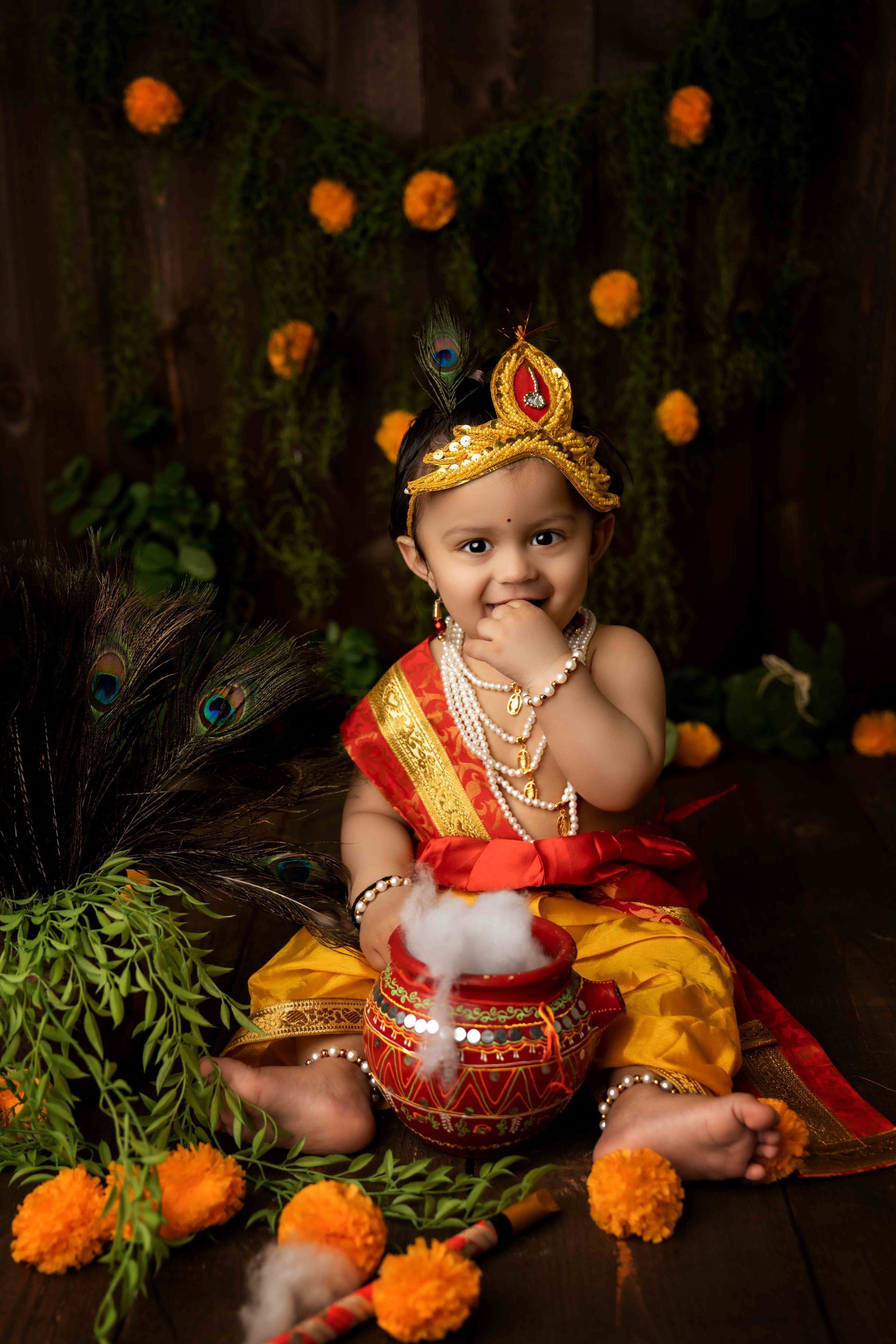 A baby in a krishna costume is sitting next to a pot.