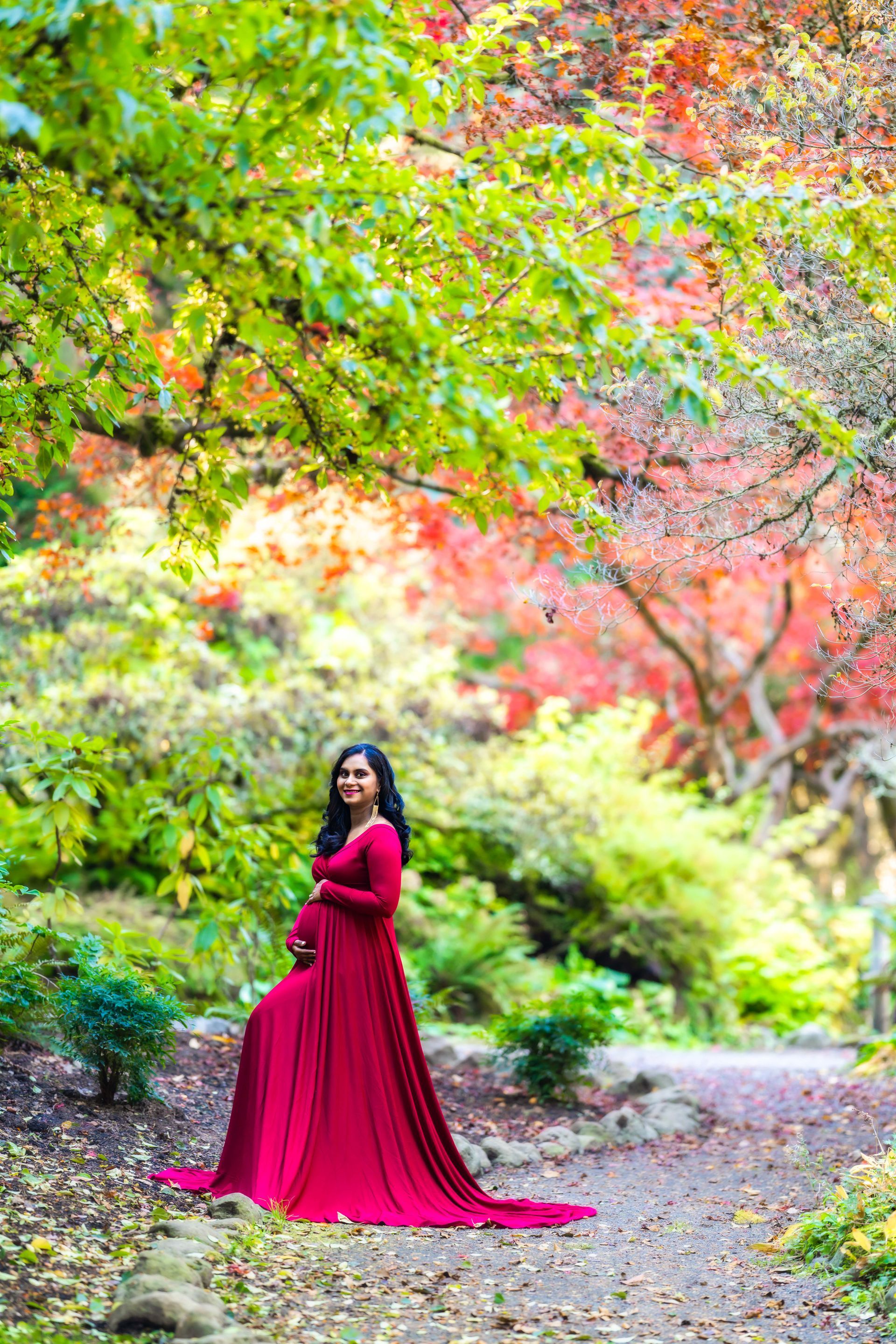 A pregnant woman in a red dress is standing in a park.