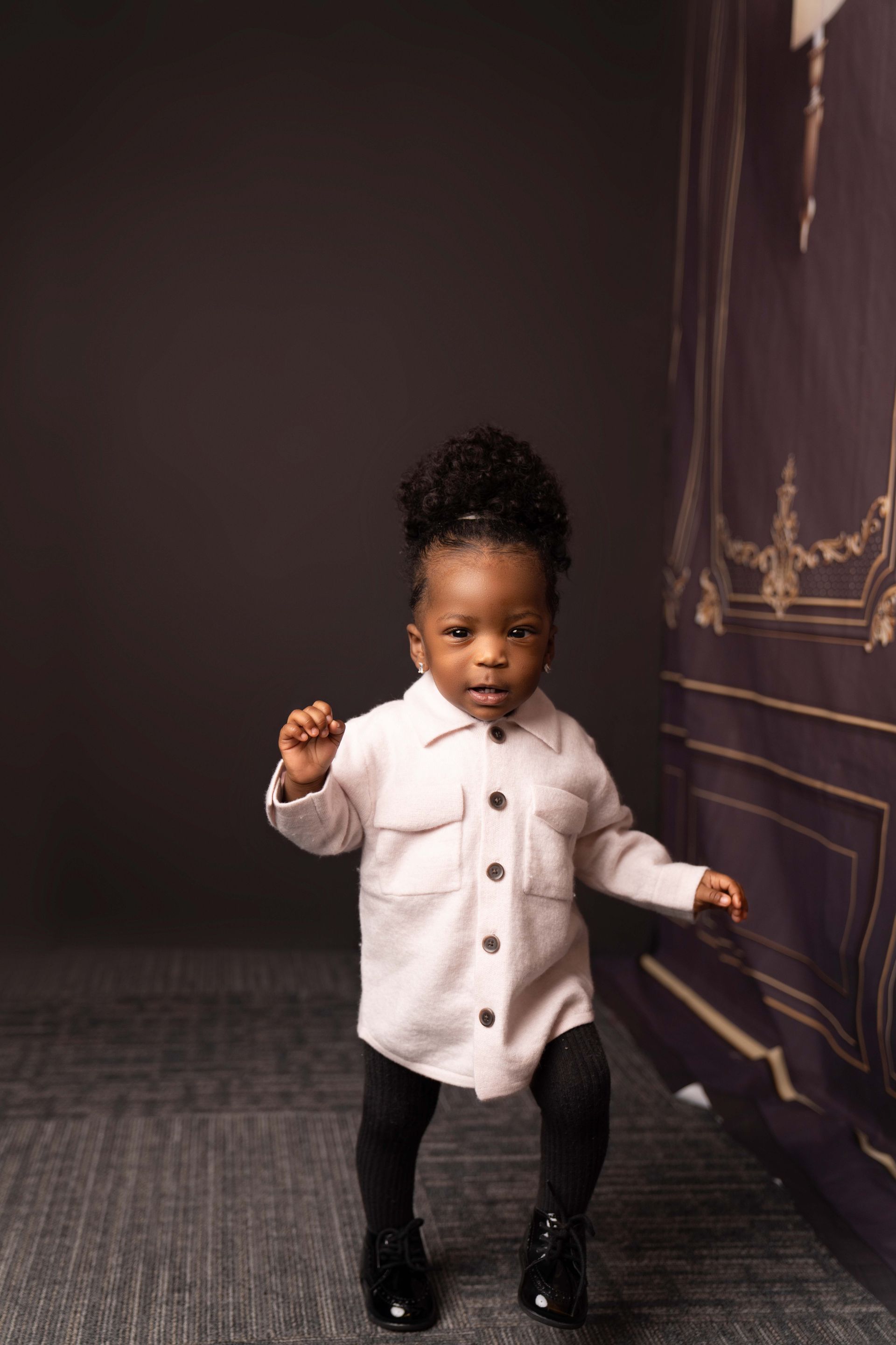 A little girl is standing in front of a wall wearing a white jacket and black pants.