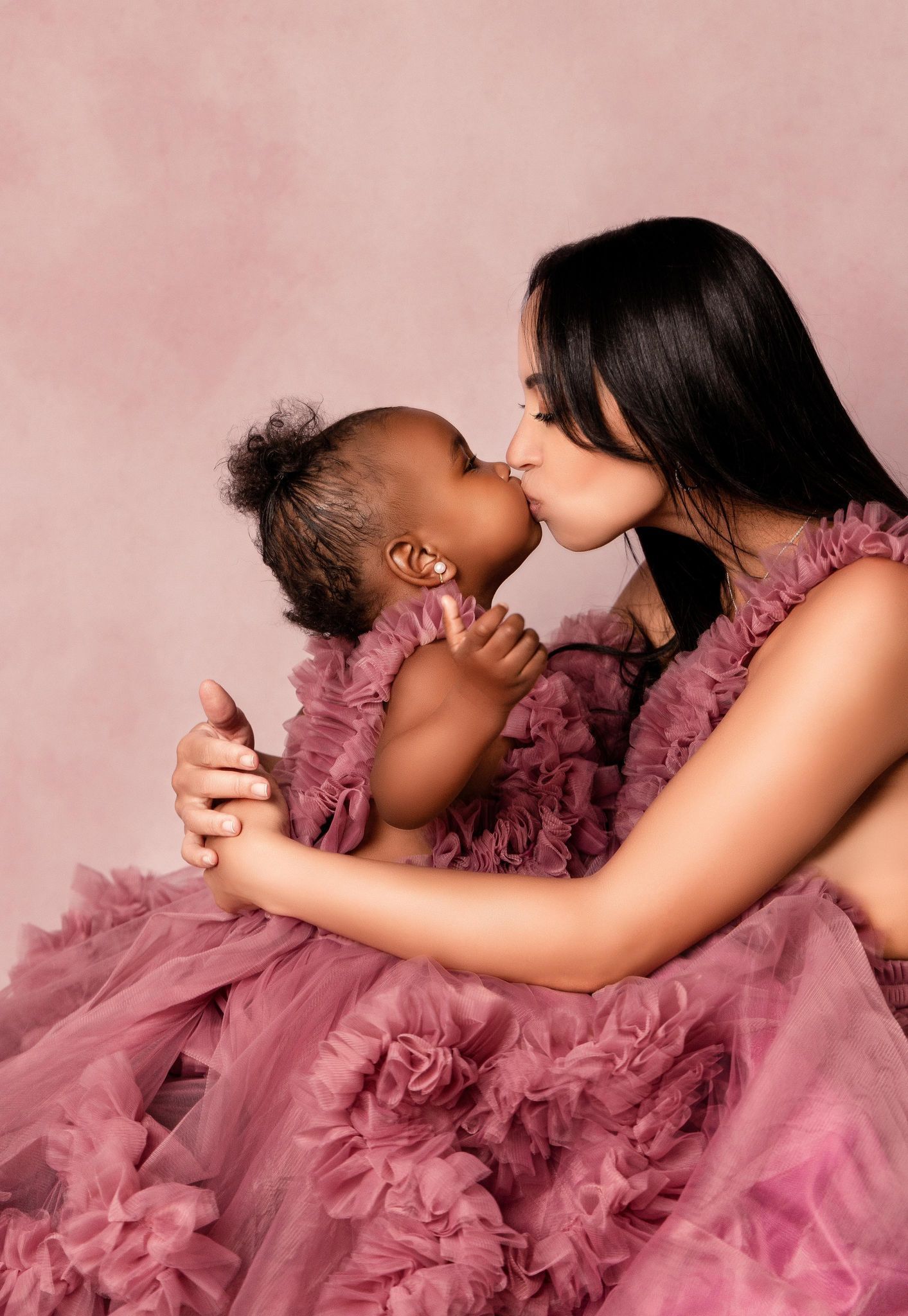 A woman in a pink dress is kissing a baby on the cheek.