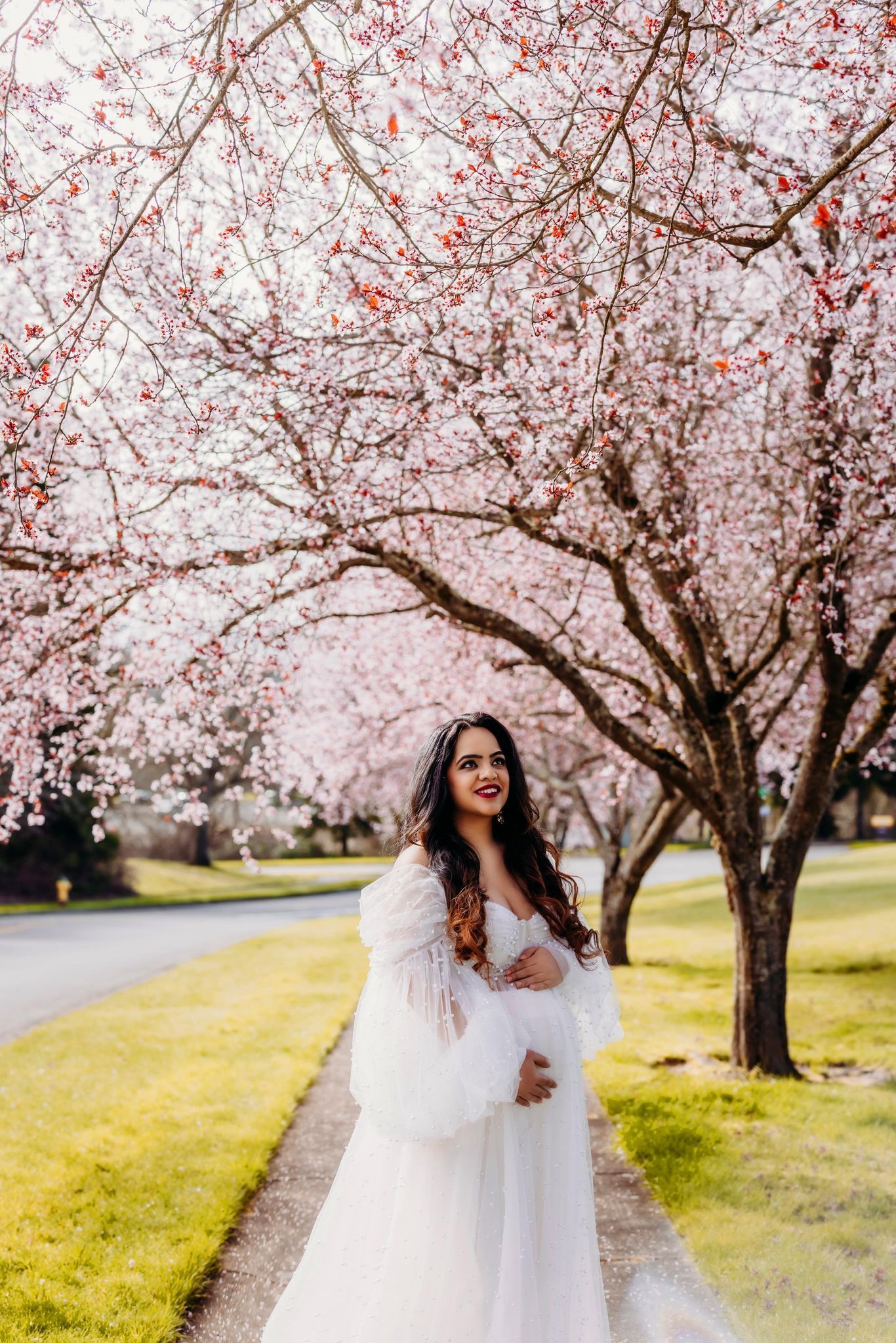 A pregnant woman in a white dress is standing under a cherry blossom tree.