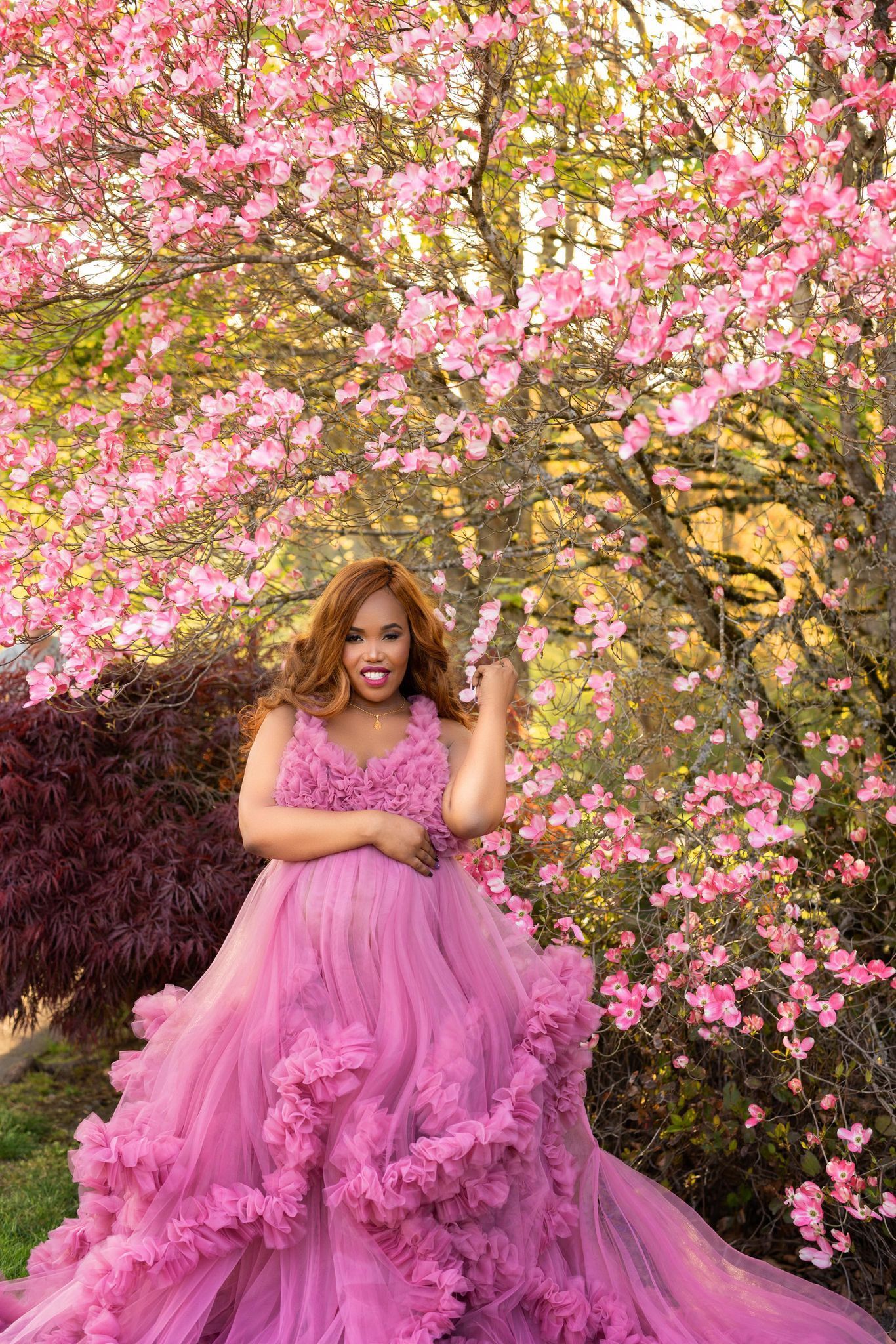 A pregnant woman in a long red dress is standing in a room surrounded by flowers.