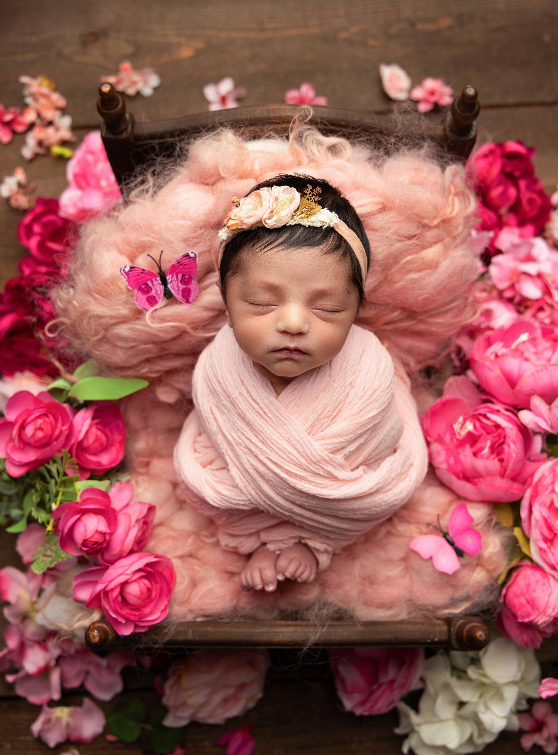 A newborn baby is wrapped in a pink blanket and surrounded by pink flowers.