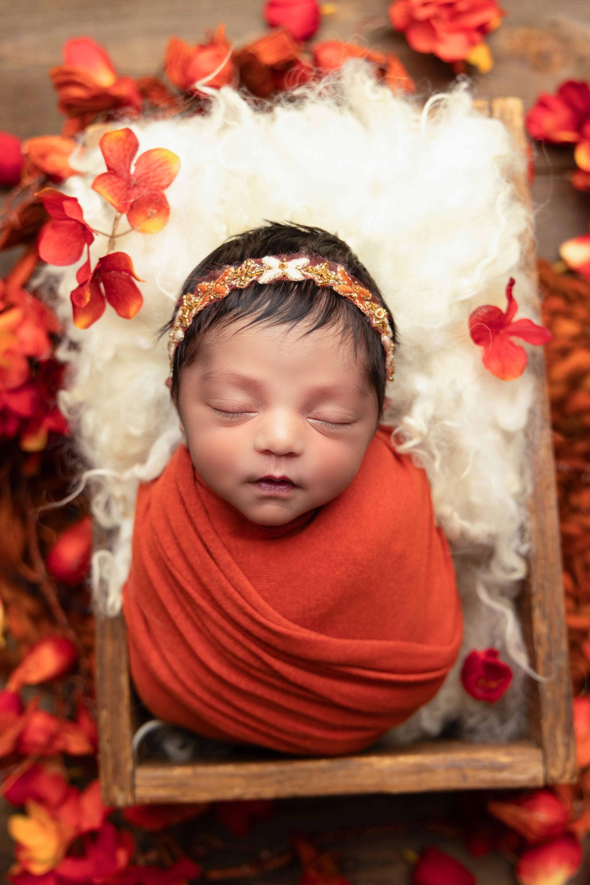 A newborn baby wrapped in an orange blanket is sleeping in a wooden box surrounded by flowers.