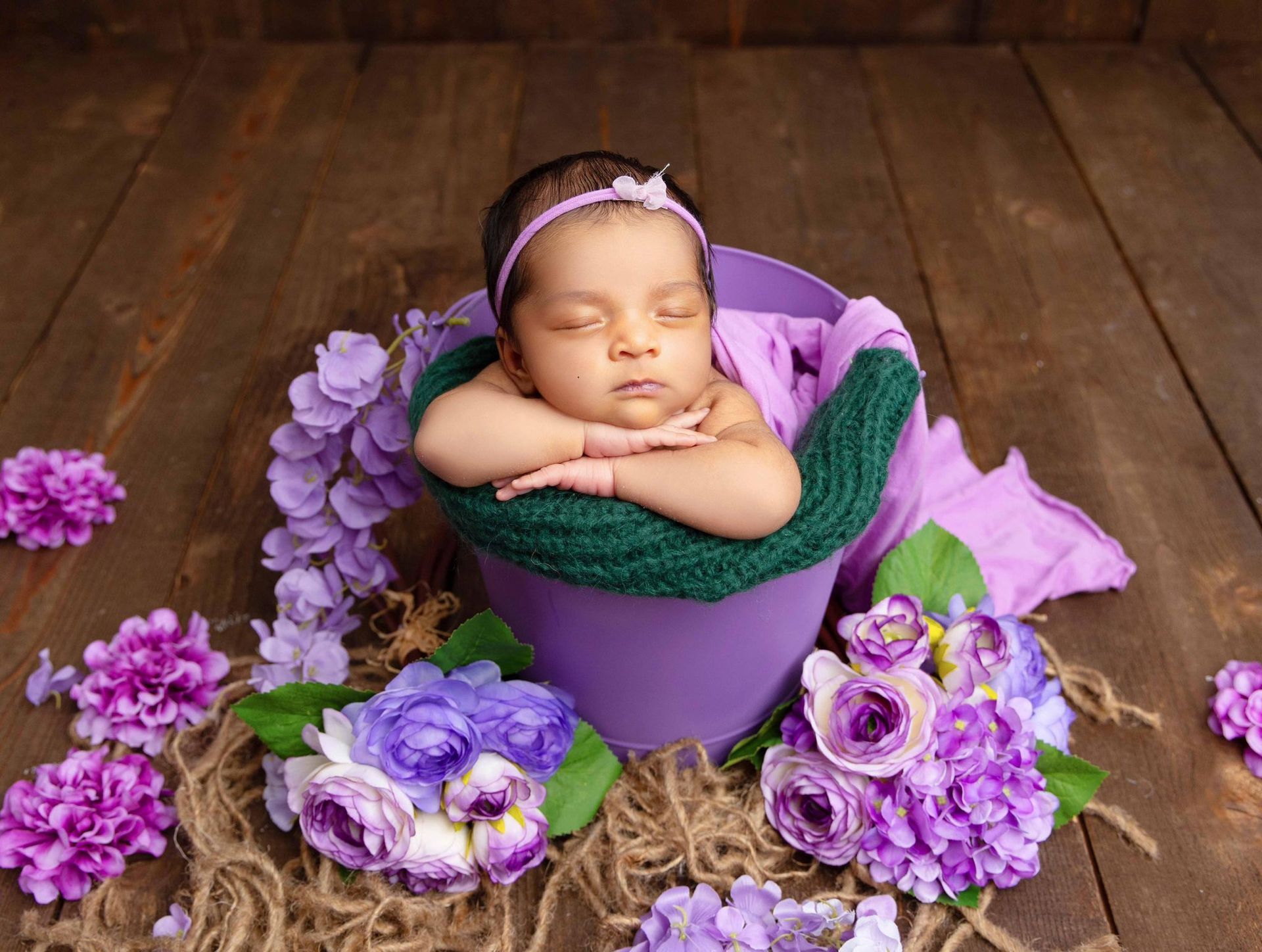 A newborn baby is sleeping in a purple bucket surrounded by purple flowers.