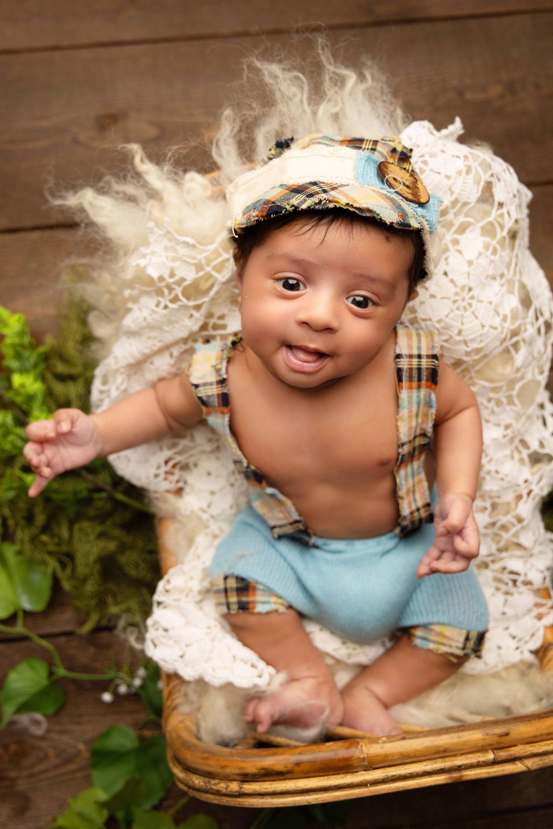A baby is sitting in a basket wearing a hat and overalls.