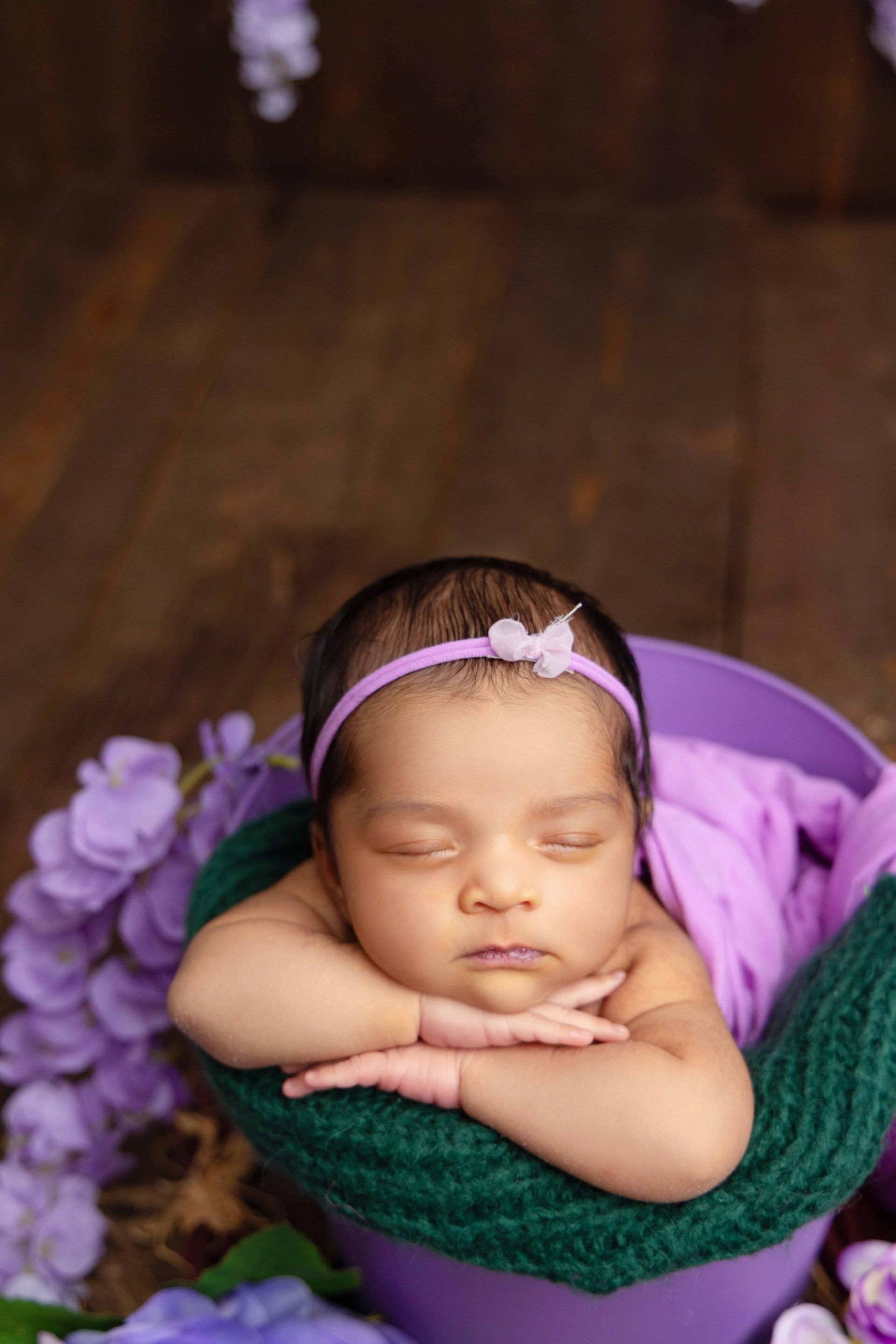 A newborn baby is sleeping in a purple bucket surrounded by purple flowers.