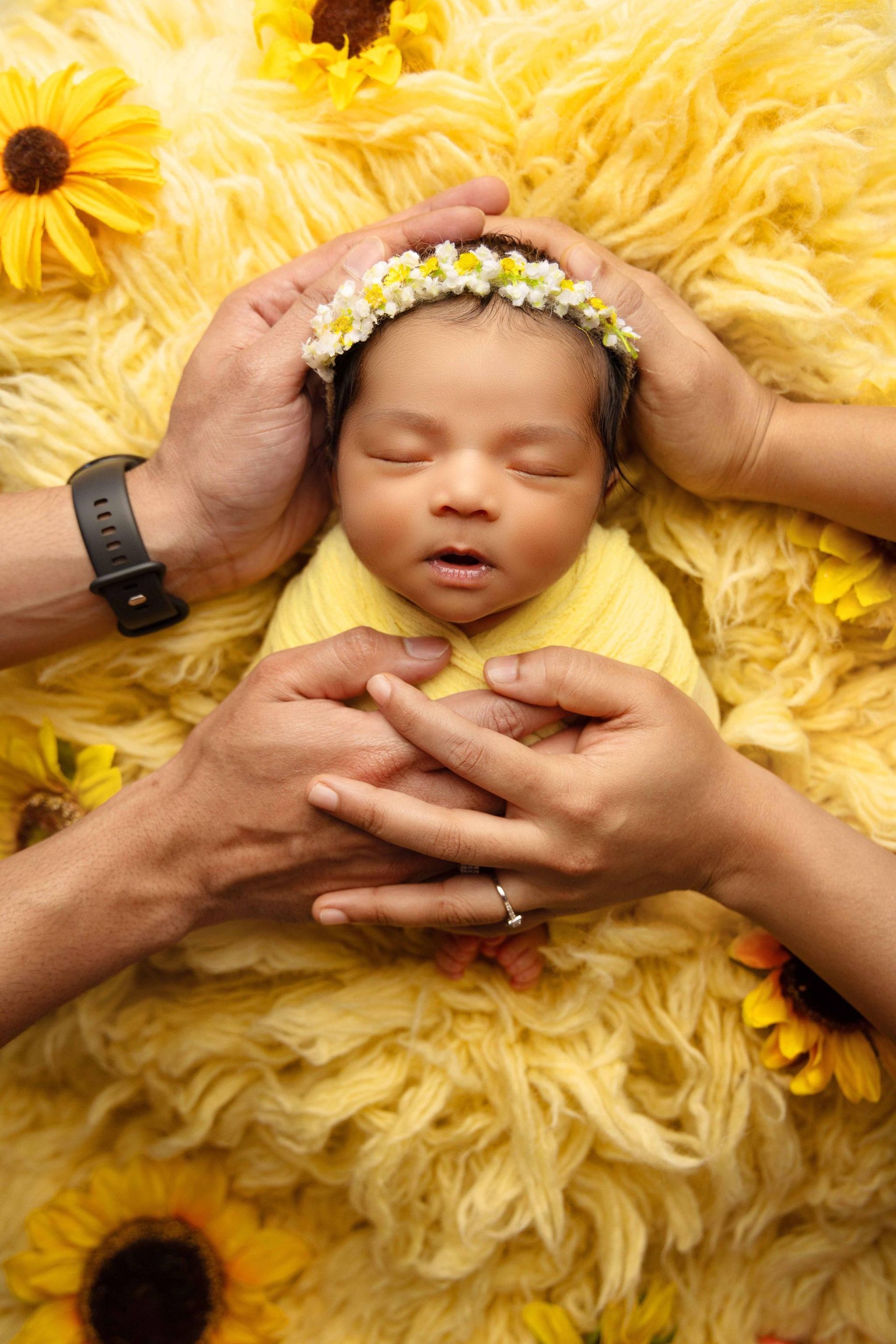 A newborn baby wrapped in a yellow blanket is laying on a yellow rug while two sets of hands hold the baby.