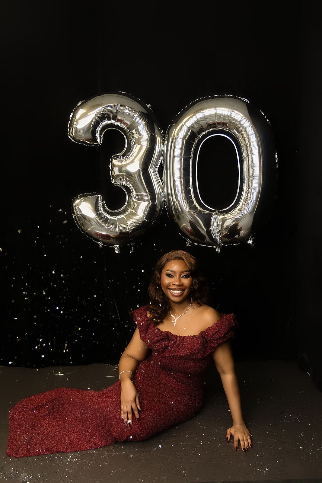 A woman in a red dress is sitting next to a large number 30 balloon.