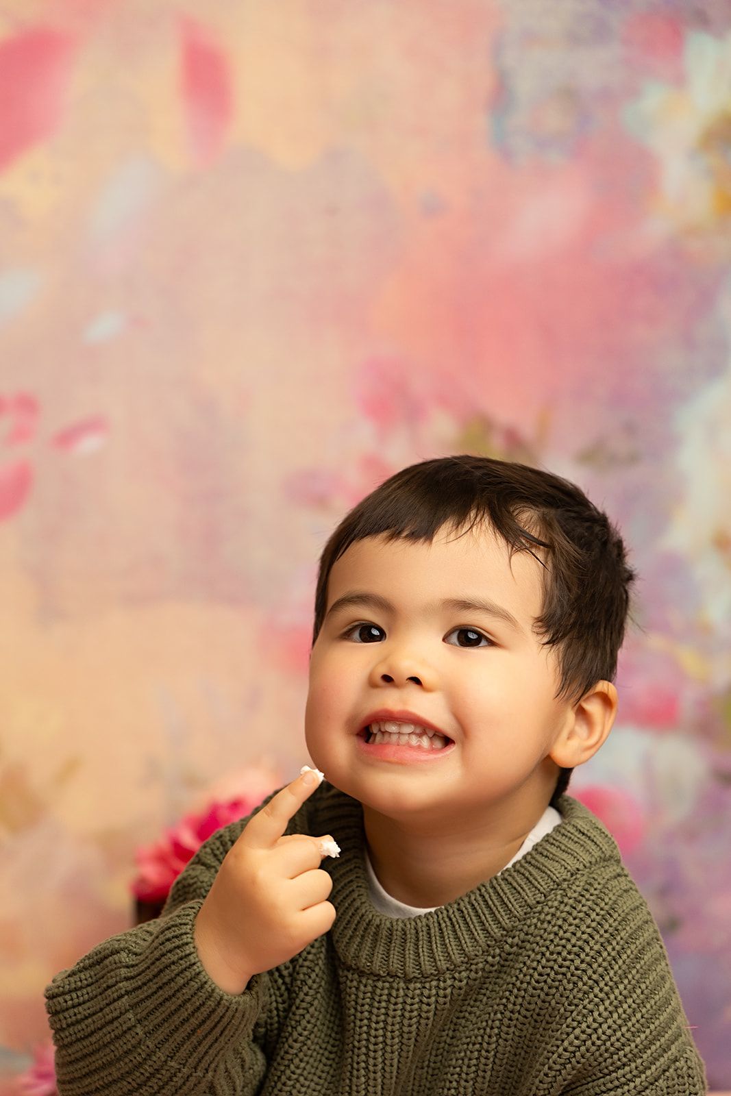 A little boy in a green sweater is pointing at his face.