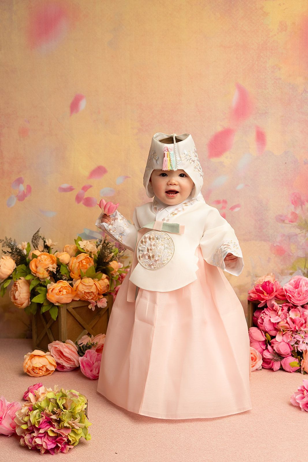 A baby in a pink dress is standing in front of flowers.