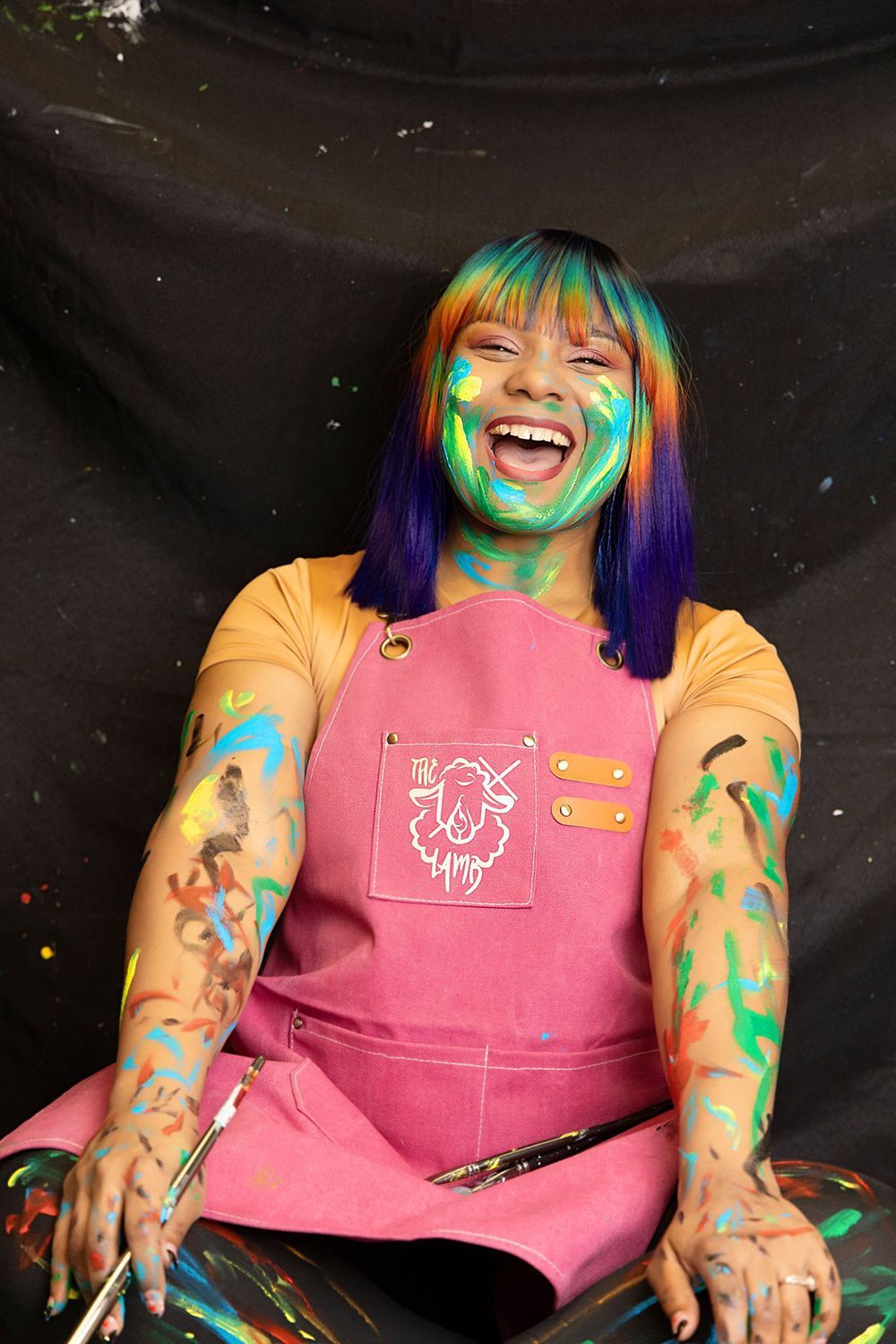 A woman with a pink apron on is covered in paint and holding a brush