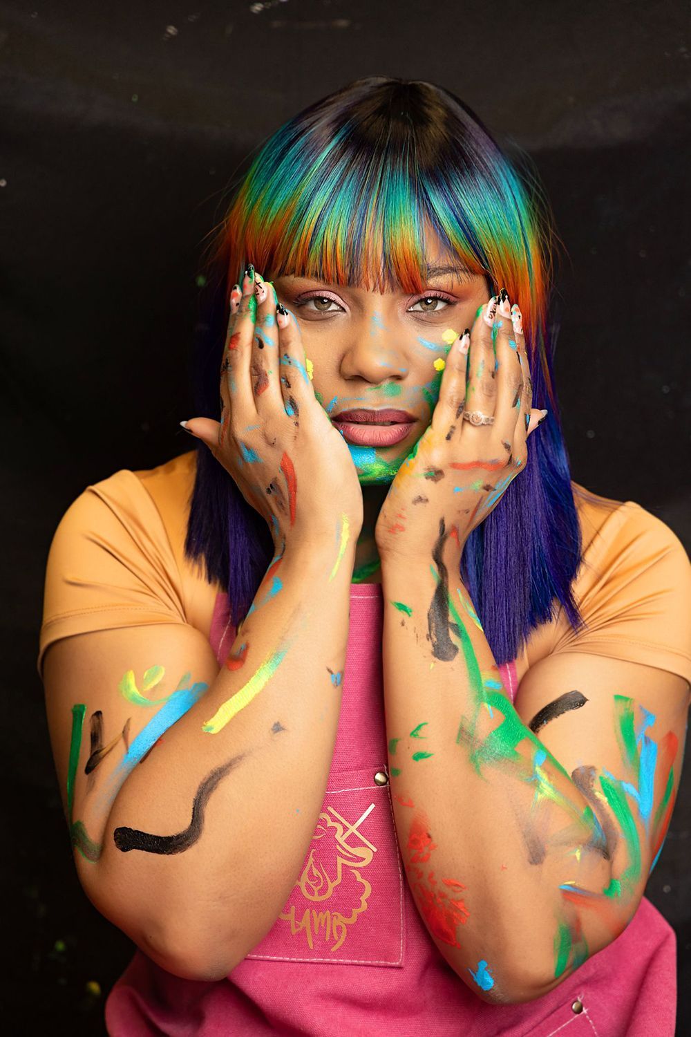 A woman with rainbow hair is covering her face with her hands covered in paint