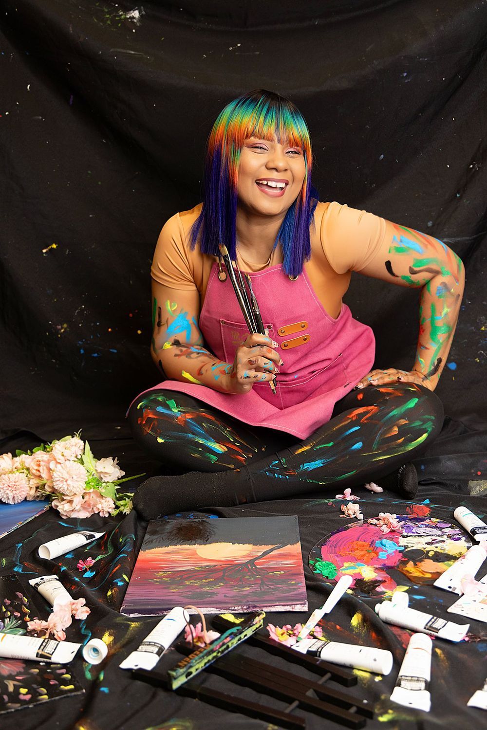 A woman with rainbow hair is sitting on the floor surrounded by paint