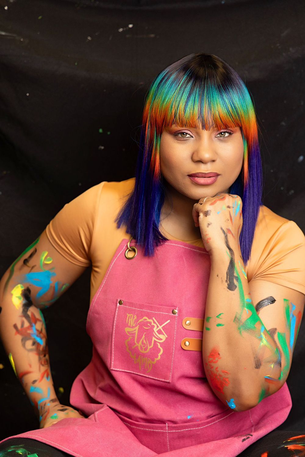 A woman with rainbow hair and a pink apron is covered in paint