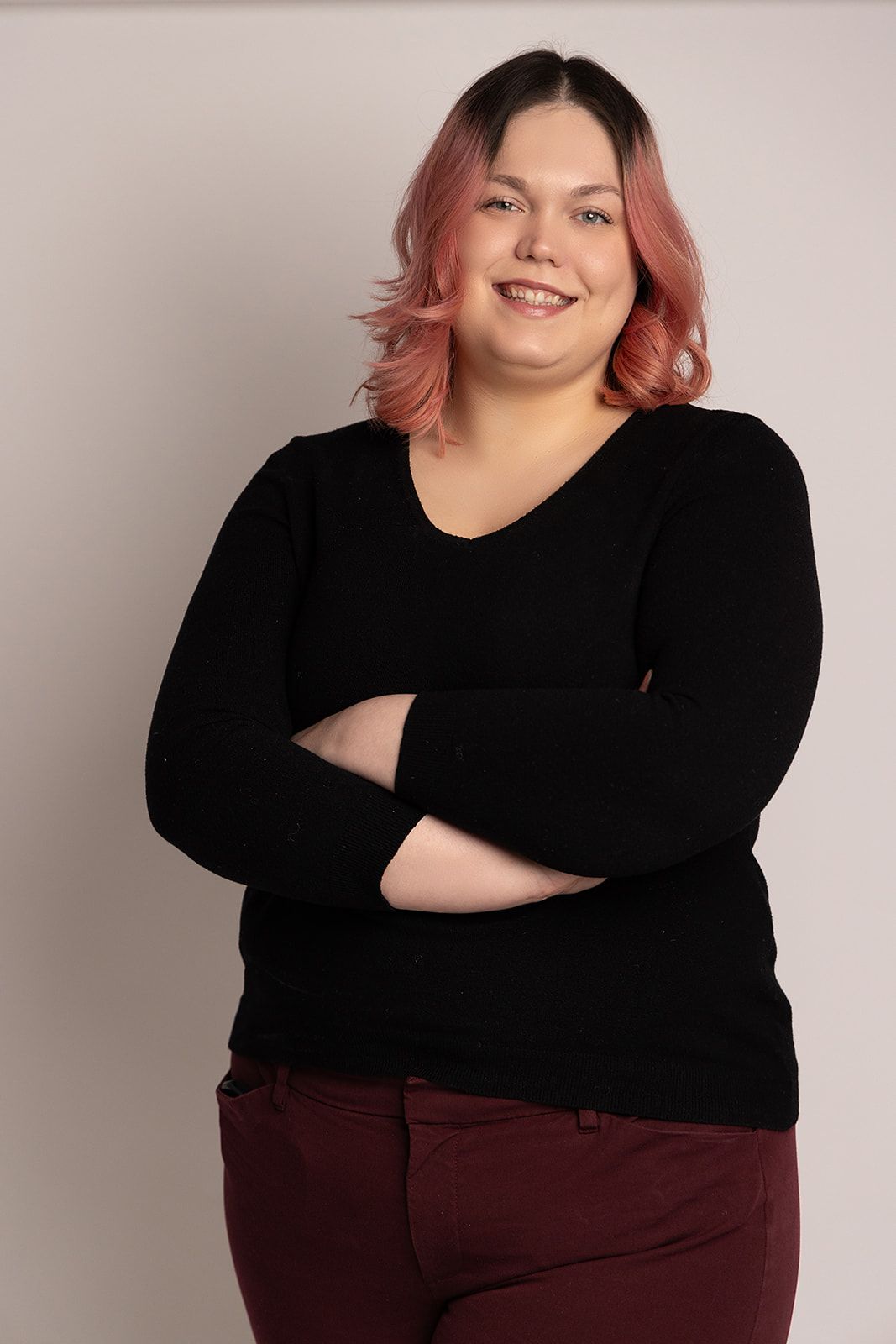 A woman with pink hair is standing with her arms crossed and smiling