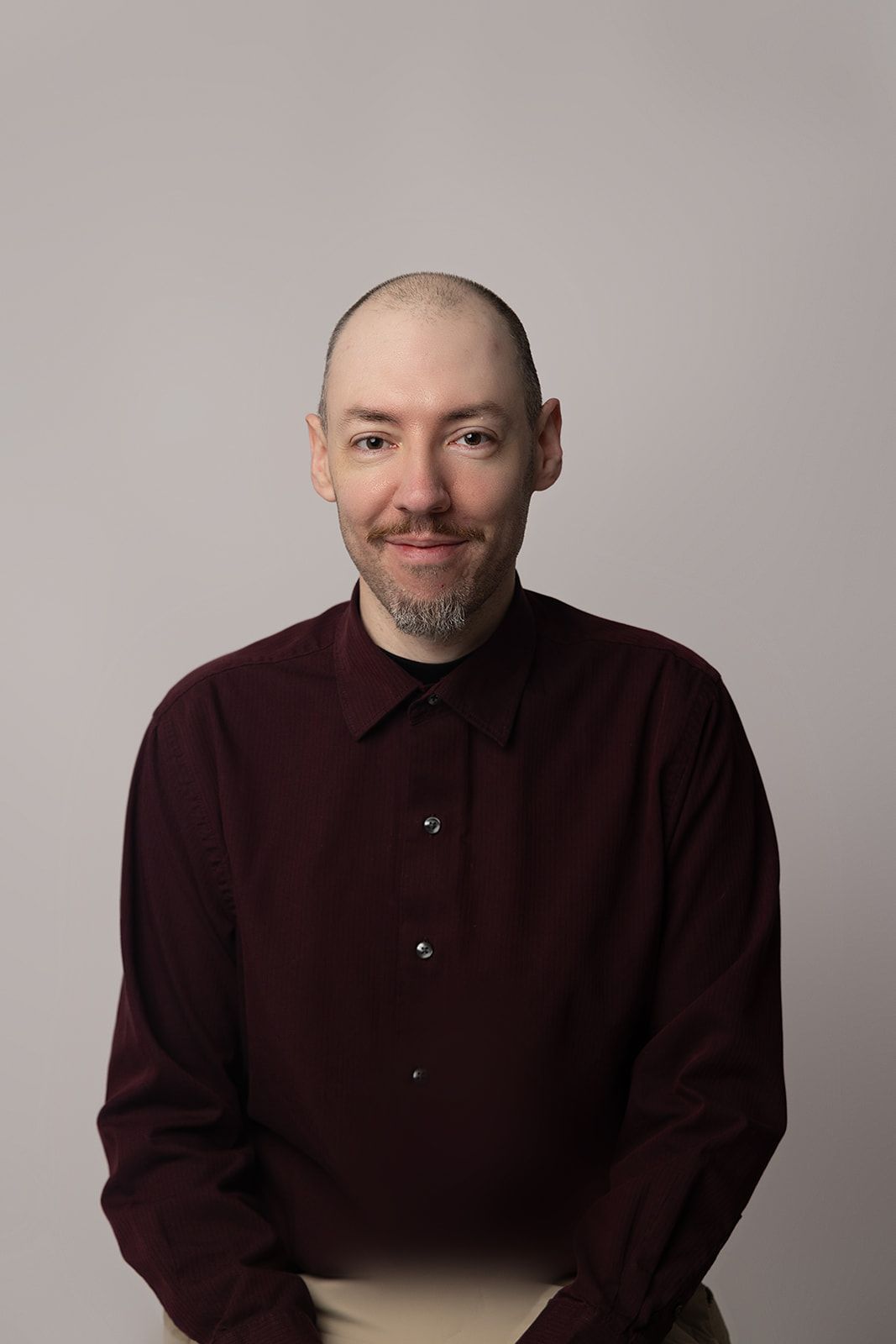 A bald man with a beard is wearing a maroon shirt and smiling
