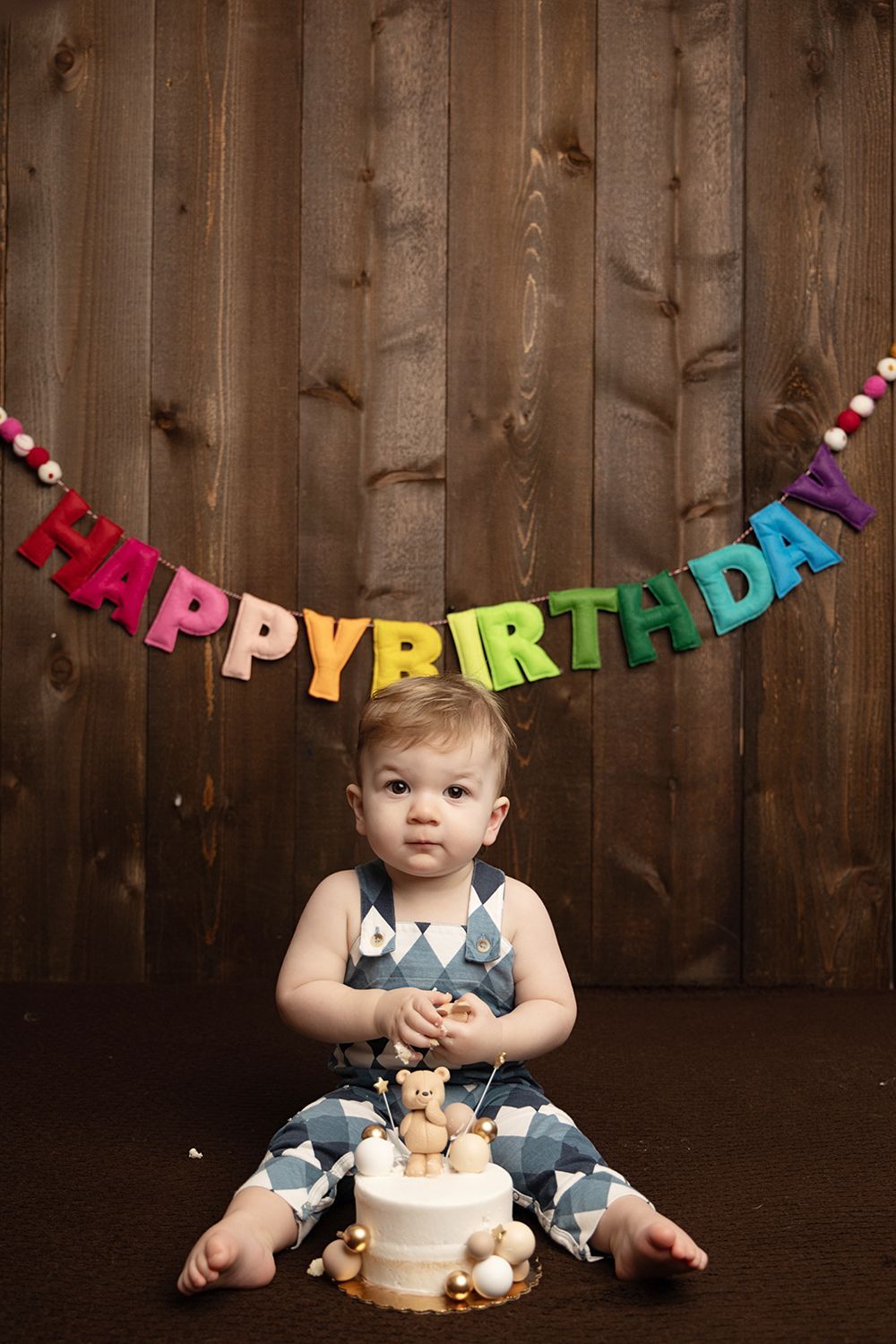 A baby is sitting next to a birthday cake and a happy birthday banner