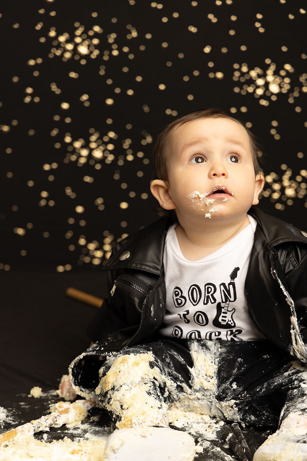 A baby in a leather jacket is sitting in a pile of cake