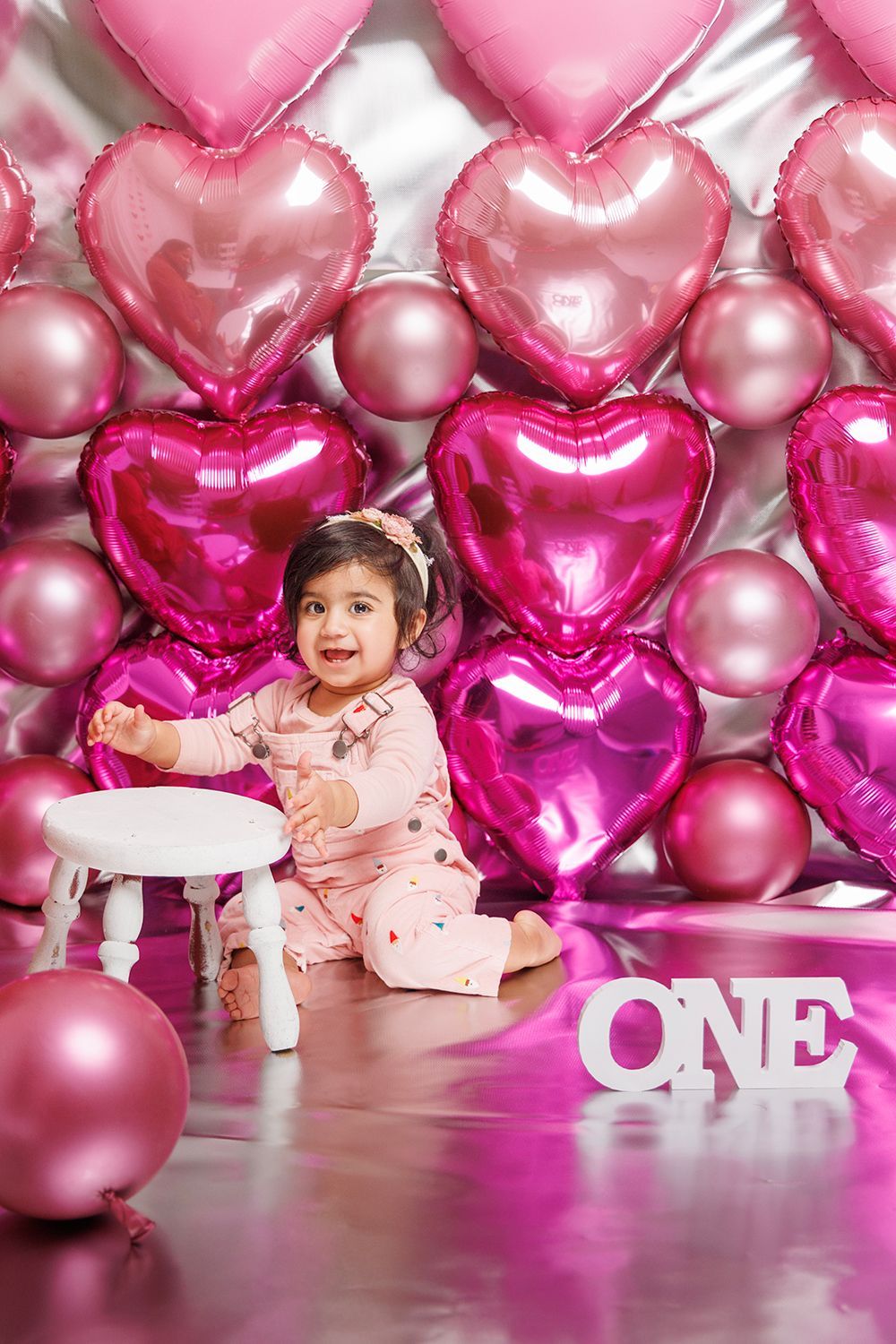 A little girl is sitting in front of a wall of pink heart-shaped balloons