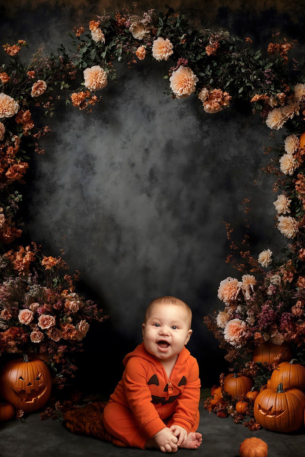 A baby in a pumpkin costume is sitting in front of pumpkins and flowers