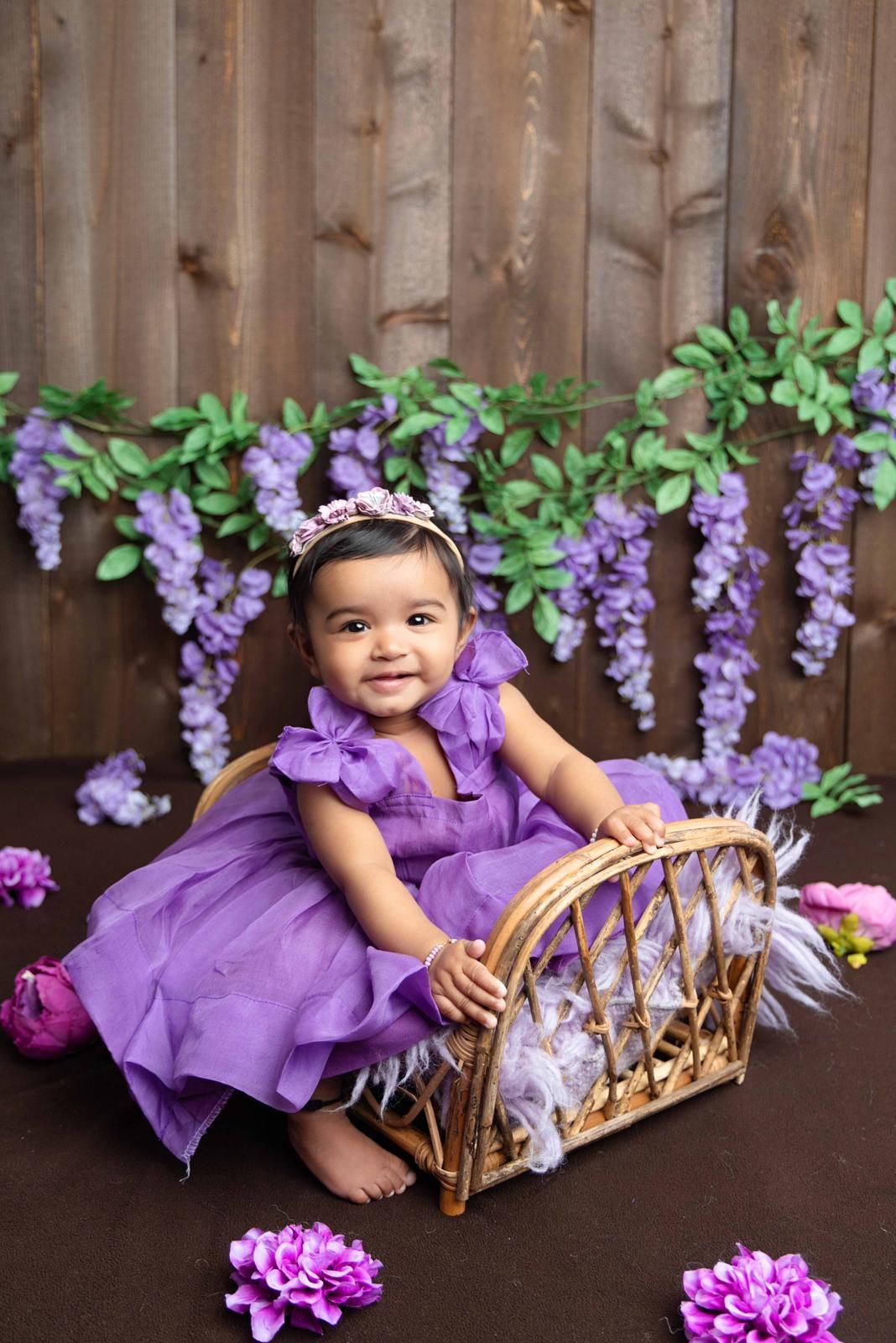 A little girl in a purple dress is sitting in a basket surrounded by purple flowers