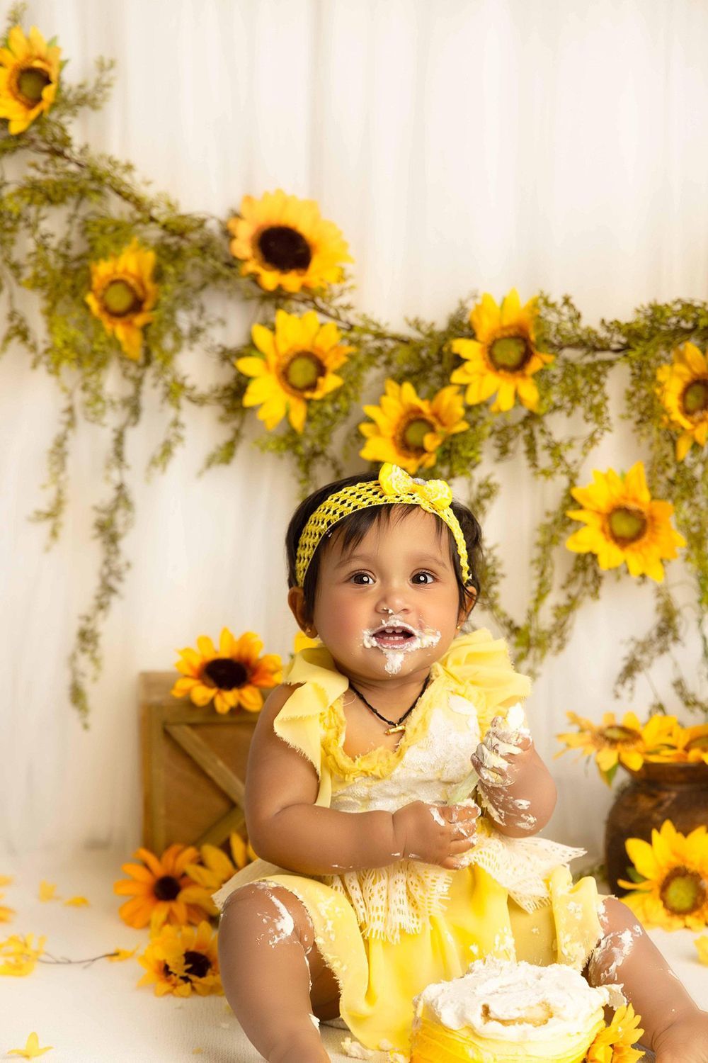 A baby girl is sitting in front of a wall of sunflowers eating a cake