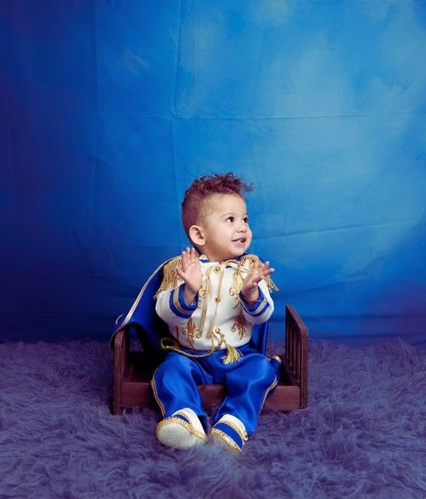 A baby dressed as a prince is sitting on a wooden chair.