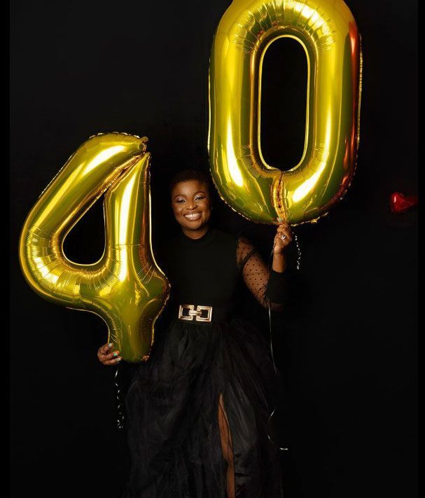 A woman in a black dress is holding two gold balloons in the shape of the number 40.