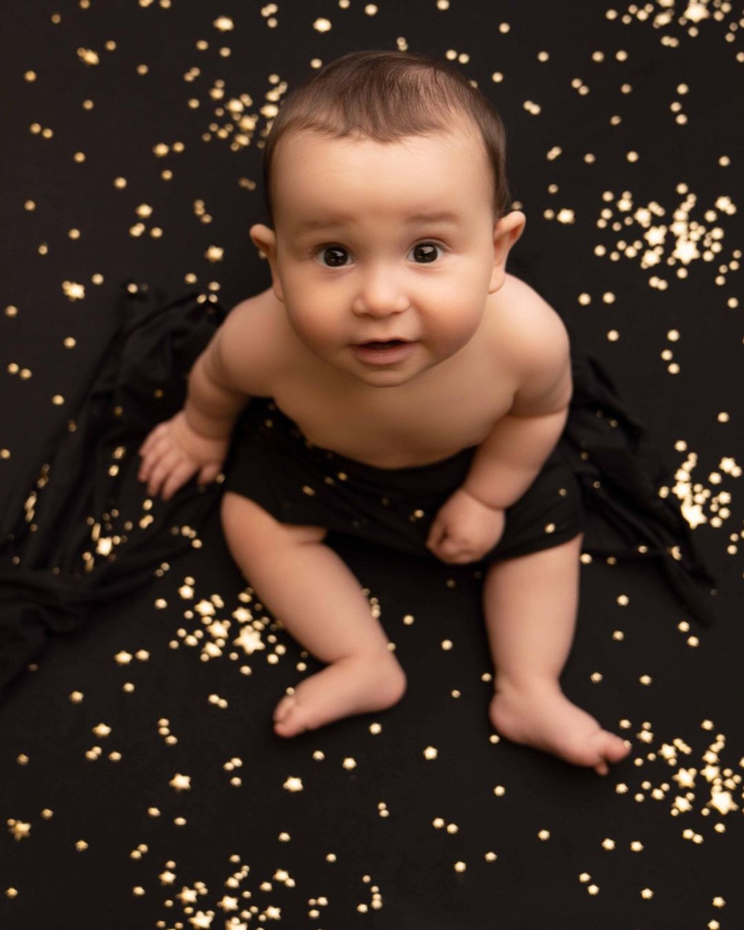 A baby is sitting on a black blanket with gold stars