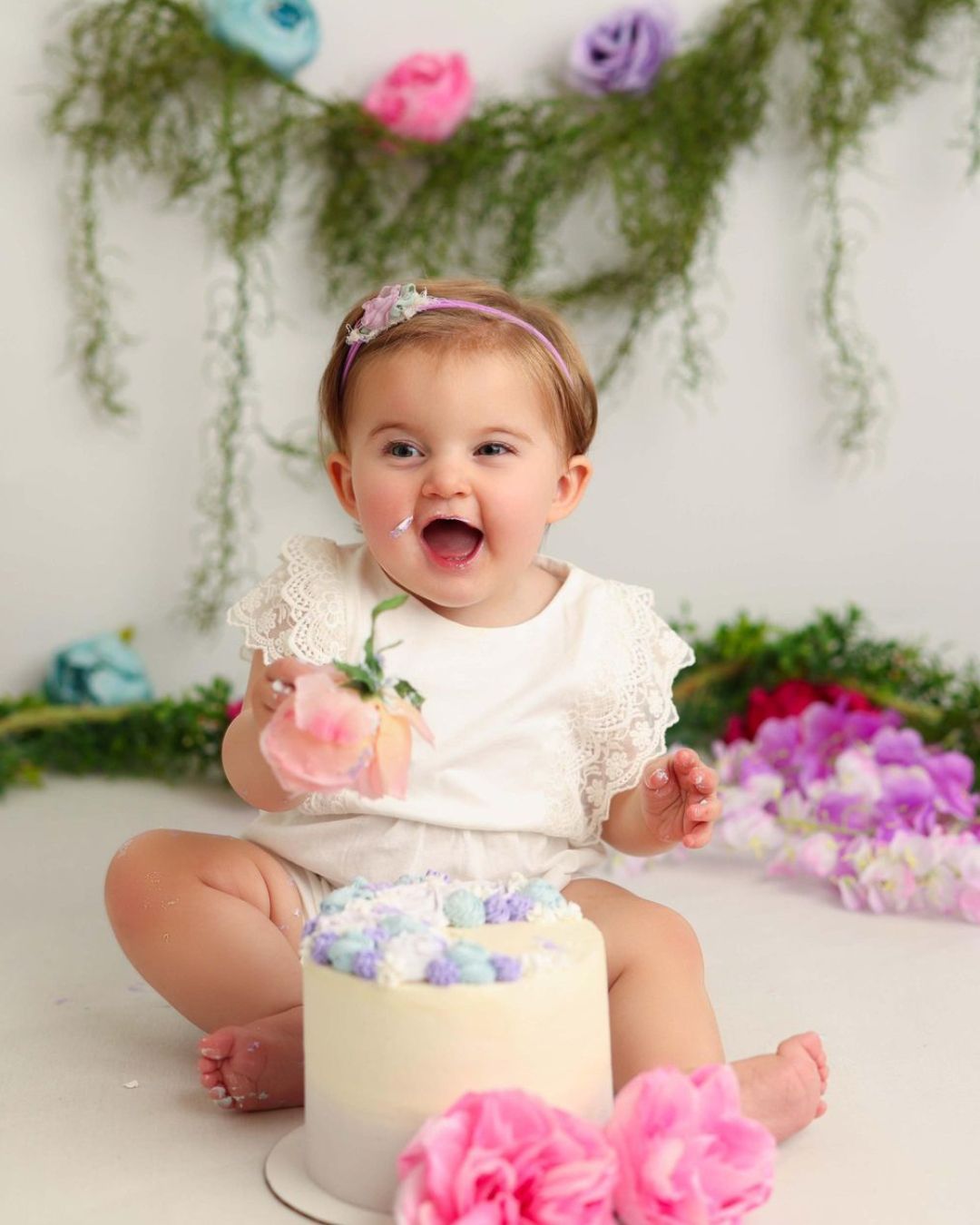 A baby girl is sitting next to a cake and flowers