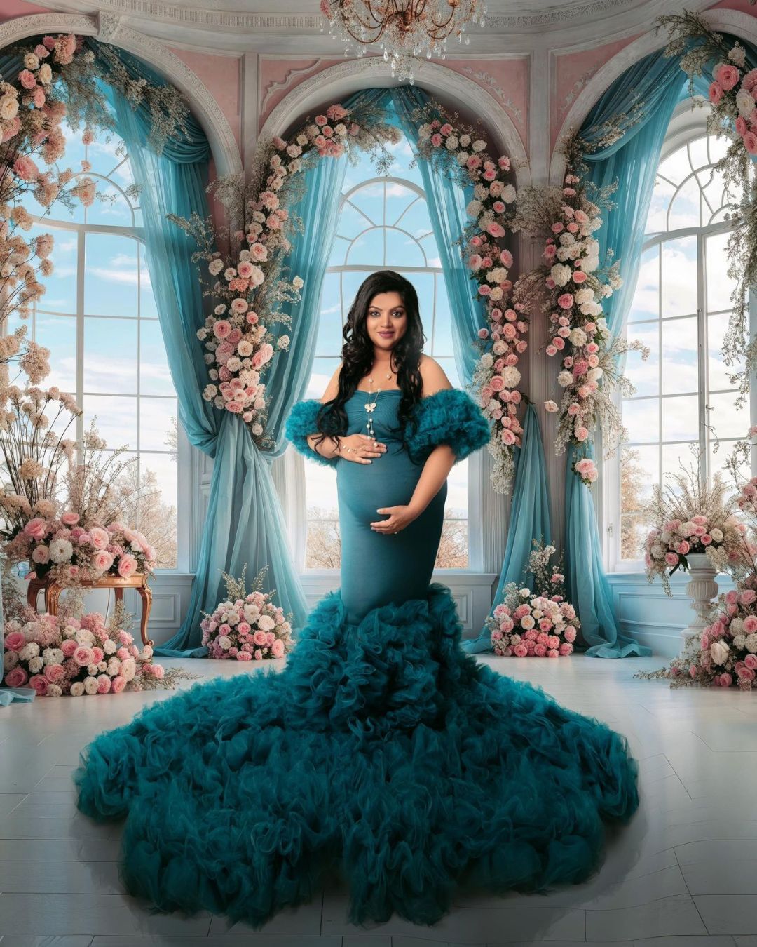 A pregnant woman in a long blue dress is standing in front of a window surrounded by flowers